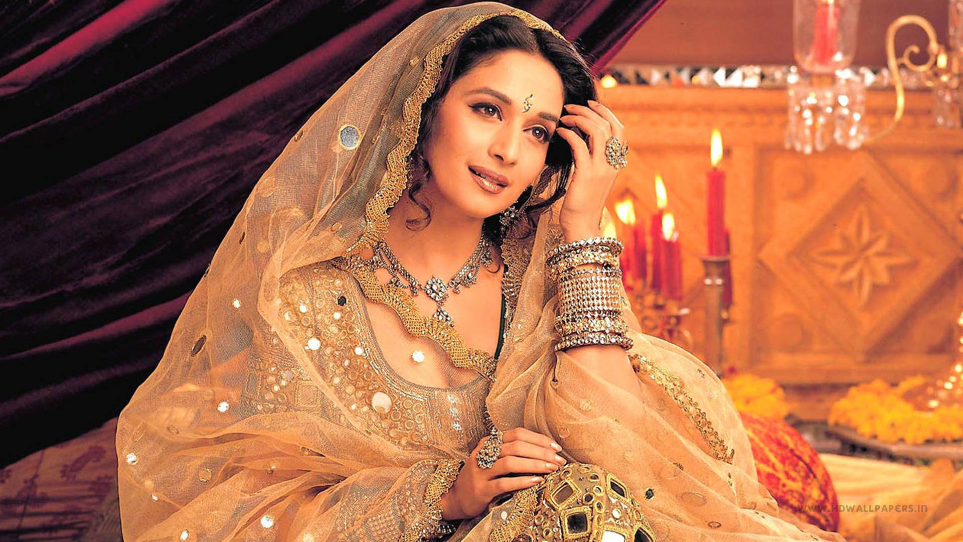 Madhuri Dixit 桌布 壁紙 — 一位身穿華麗金色傳統服飾、戴著薄紗的女性在點燃的蠟燭和花卉供品旁溫柔微笑。