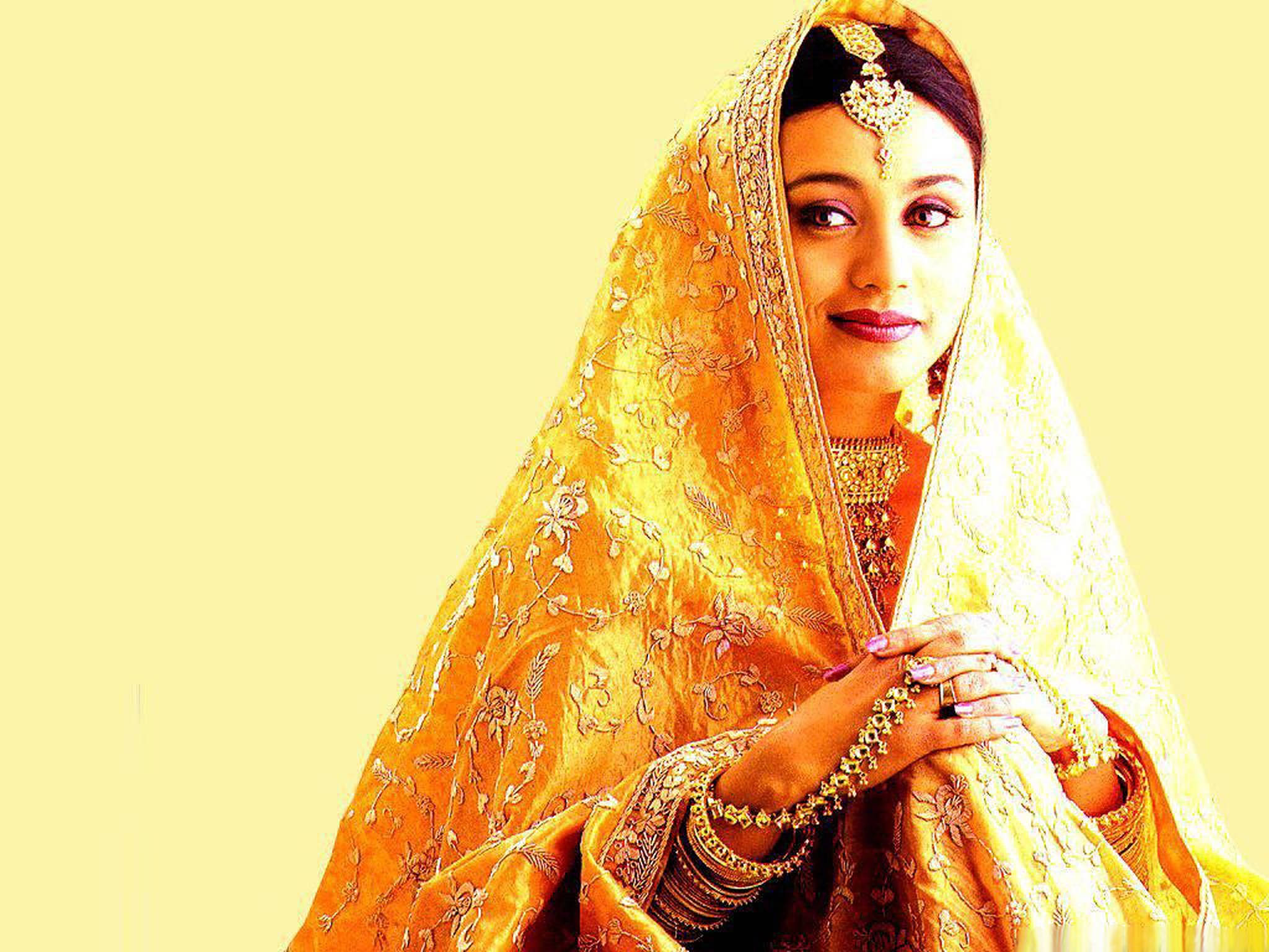 Rani Mukerji 桌布 壁紙 — 一位身穿華麗金色婚紗、飾有精緻刺繡和珠寶的女性，在淡黃色背景下溫柔凝視。