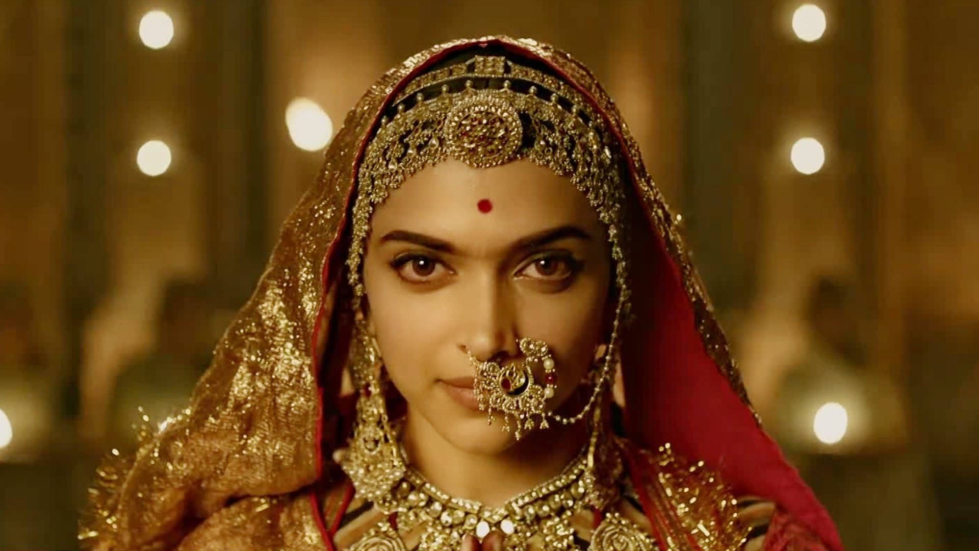 Deepika 桌布 壁紙 — 一位身穿華麗金色新娘禮服、戴著紅色頭紗和精緻珠寶的女性，在溫暖閃爍的燈光下向前凝視。