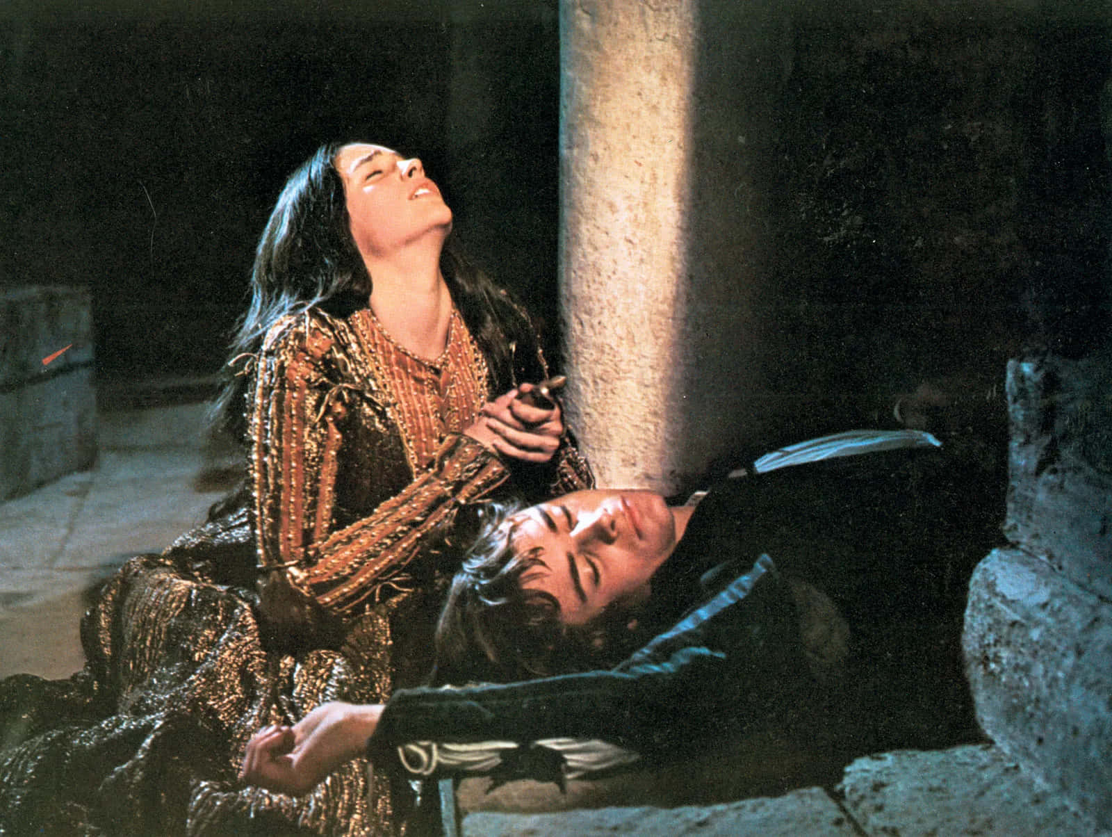 romeo and juliet 桌布 壁紙 — 一位身穿華麗金色服飾的女子，在光線昏暗的古代廢墟中，抬頭凝視著旁邊一名靠著石柱一動不動躺著的男子。