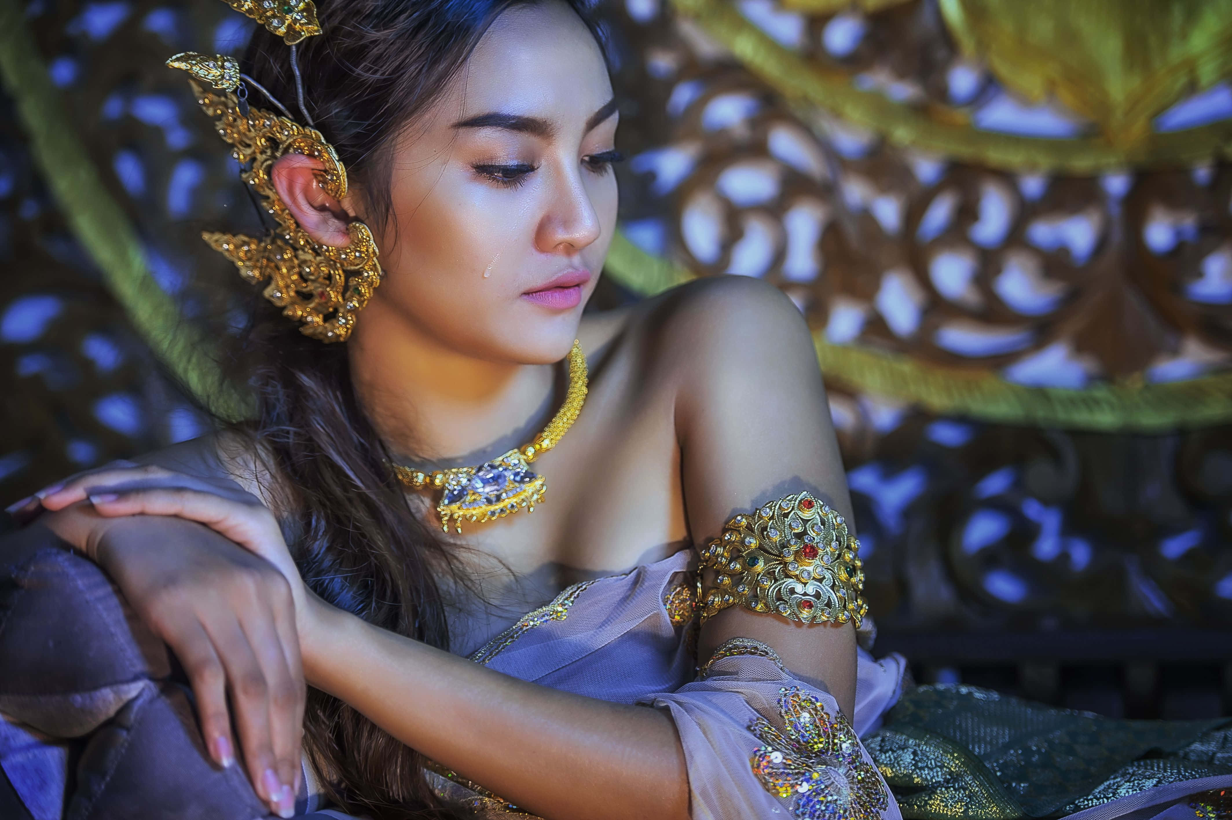 美女 桌布 壁紙 — 一位身穿華麗金色泰式珠寶和薰衣草服飾的女性，倚靠在雕刻木質背景上沉思。