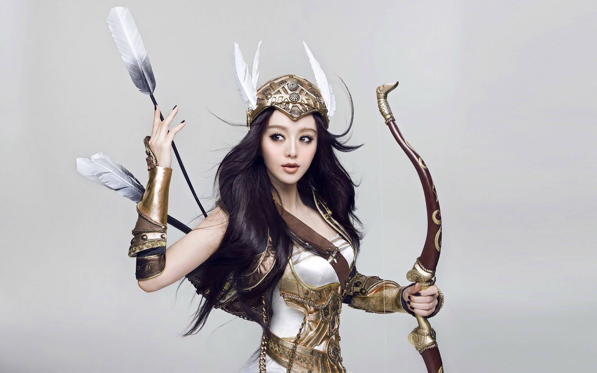 美女 桌布 壁紙 — 一位身穿華麗金色盔甲和羽毛頭盔的女性，在純灰色背景前手持弓箭。
