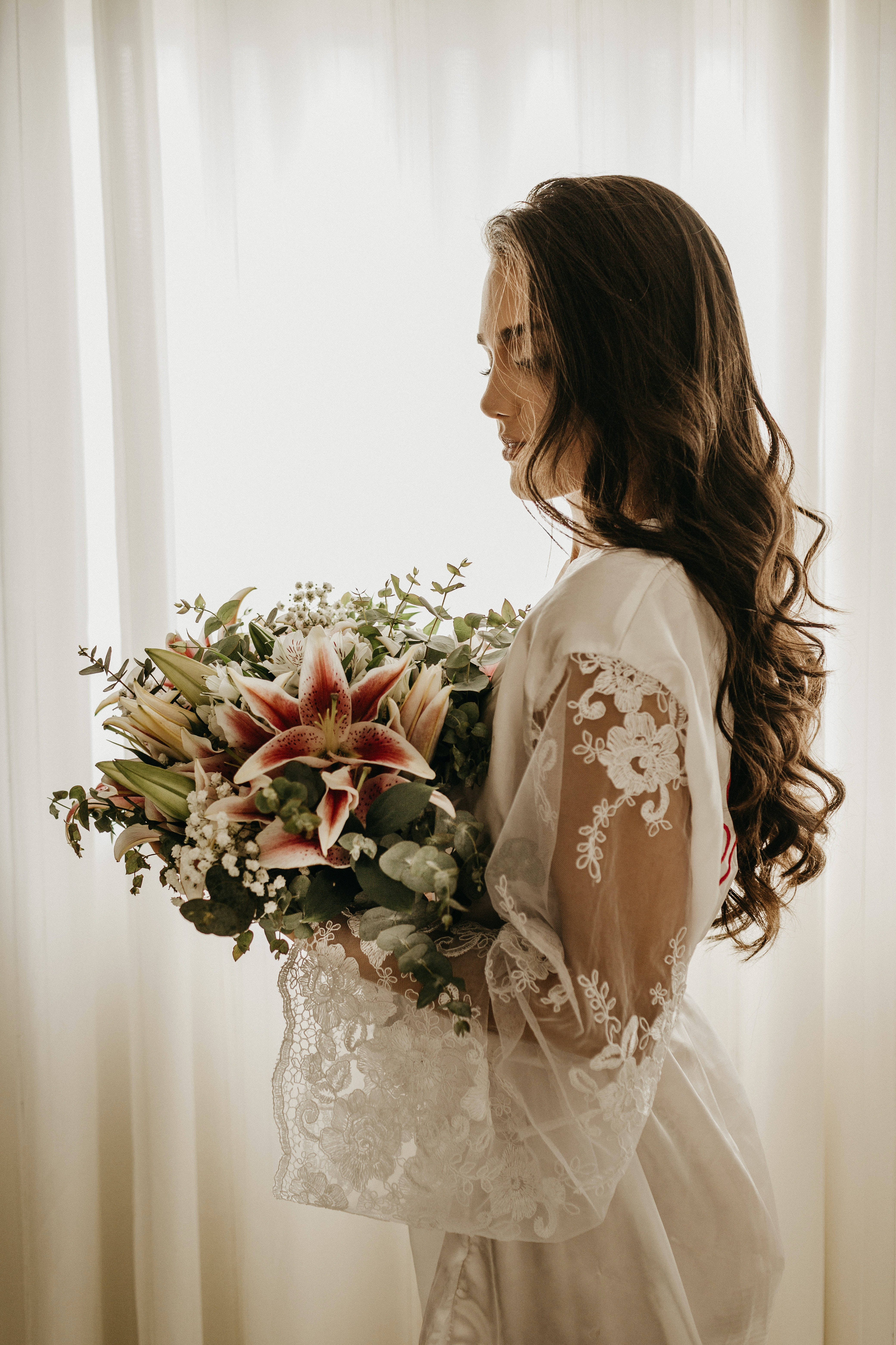 Bridal 桌布 壁紙 — 一位身穿蕾絲婚紗的新娘在陽光照射的窗邊手持一束充滿活力的百合花和綠葉花束。