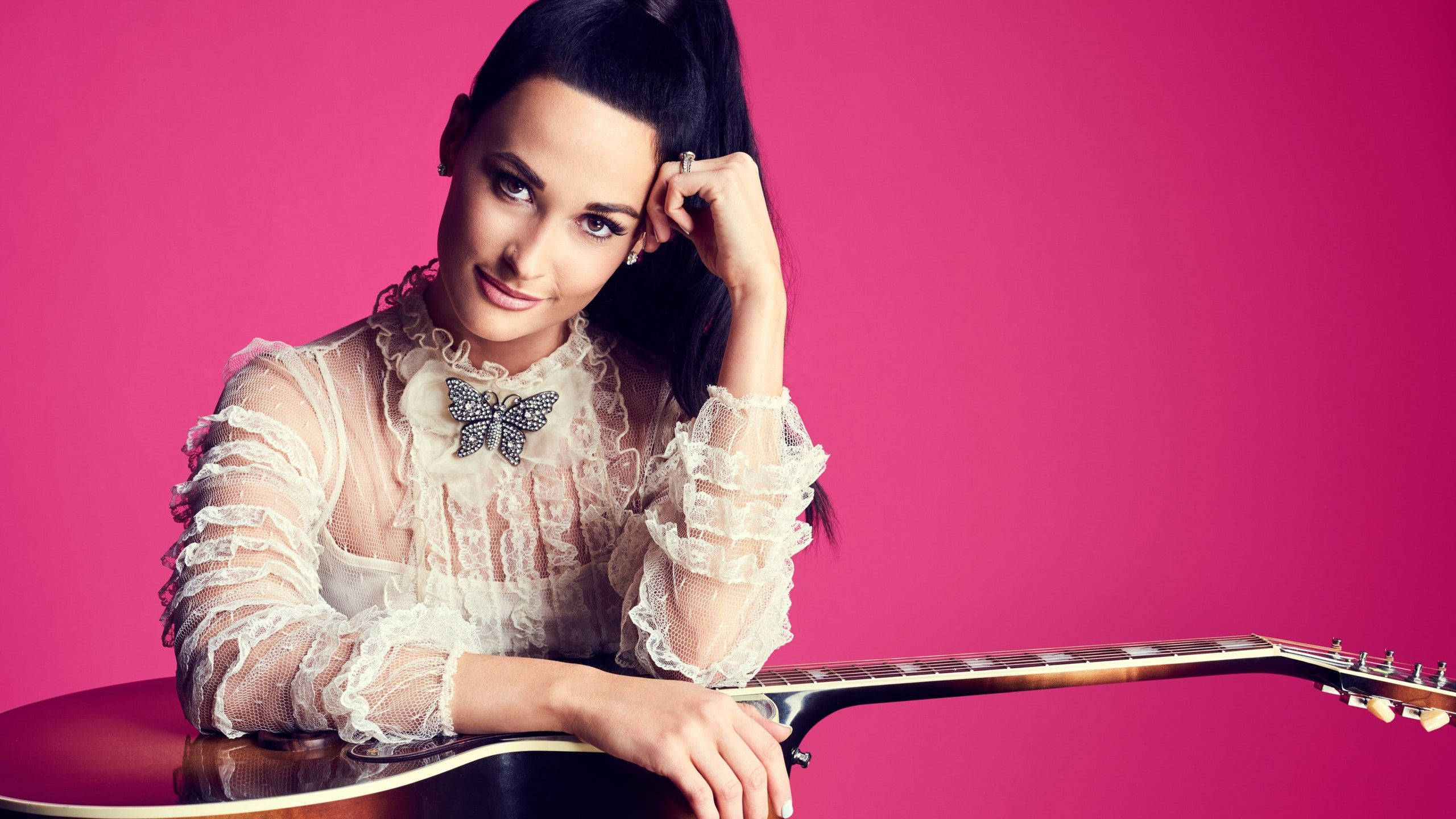 Kacey Musgraves 桌布 壁紙 — 一位身穿蕾絲襯衫、佩戴蝴蝶胸針的女性，在充滿活力的粉紅色背景前倚靠著吉他。