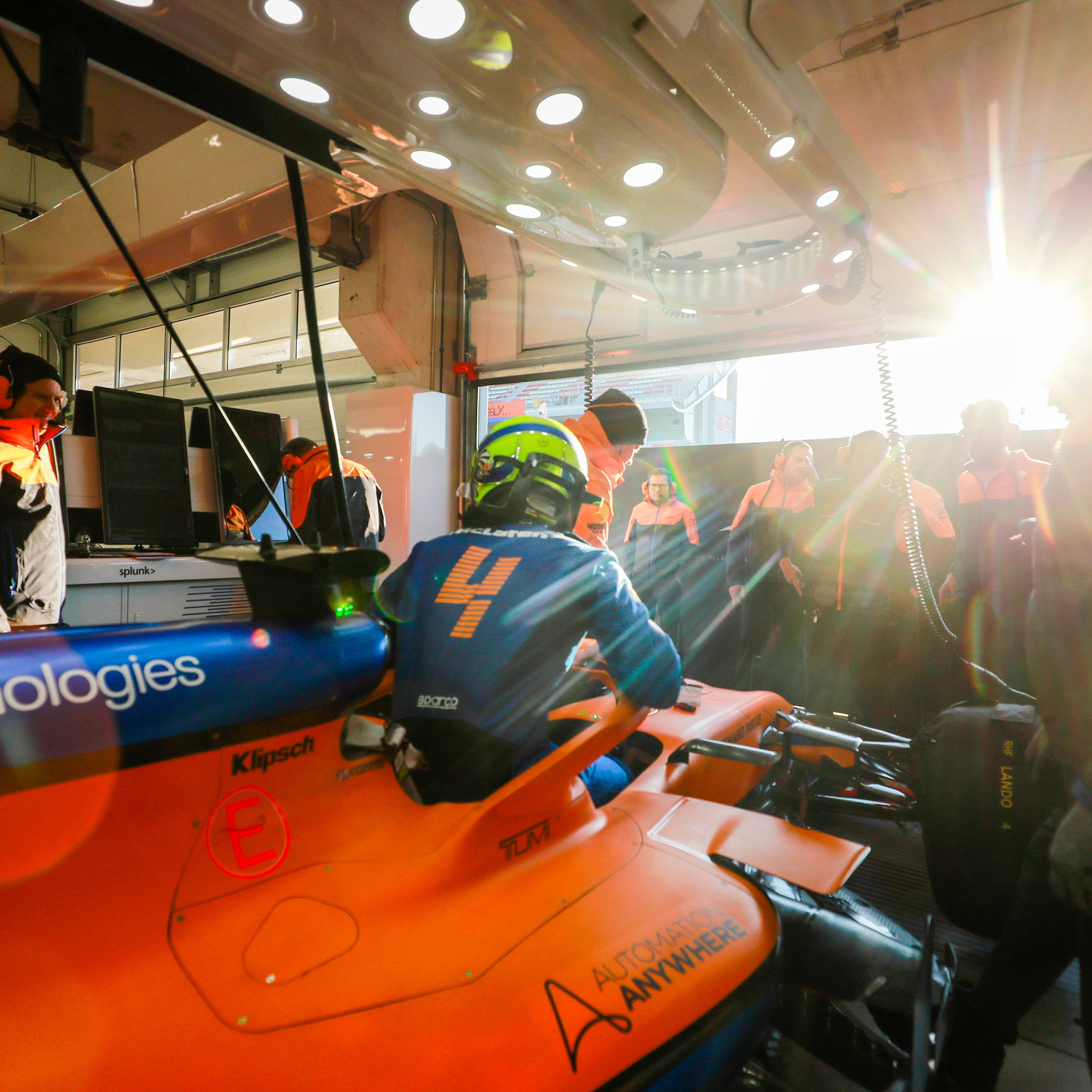 Racing 桌布 壁紙 — 一位身穿藍橙色賽車服的F1車手坐在他亮橙色的賽車中，機械師和車隊工作人員在陽光普照的車庫裡圍繞他工作。