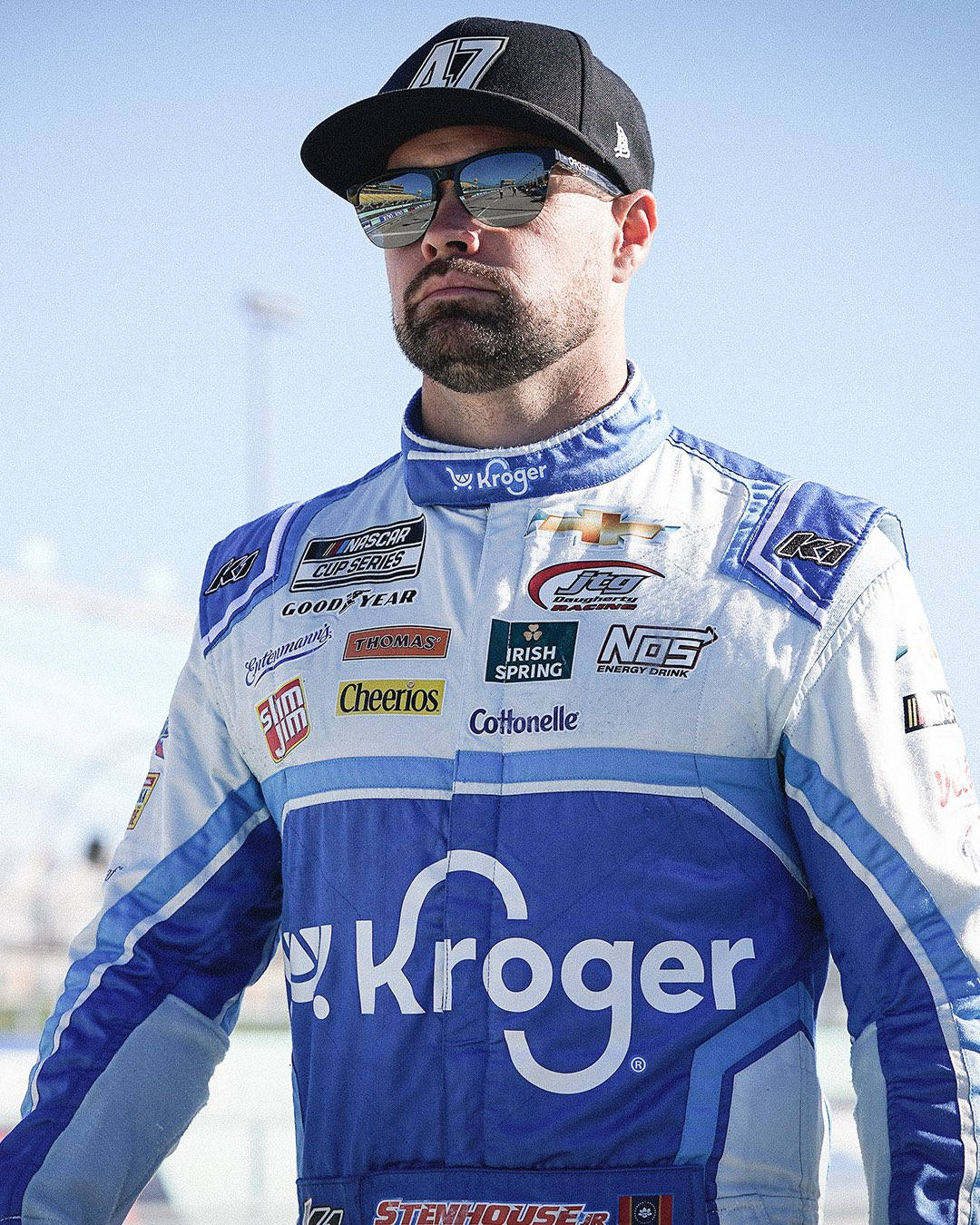 人像動態 桌布 壁紙 — 一位身穿藍白 Kroger 賽車服的 NASCAR 車手，戴著太陽眼鏡和帽子，在明亮的日光下自信地站立。