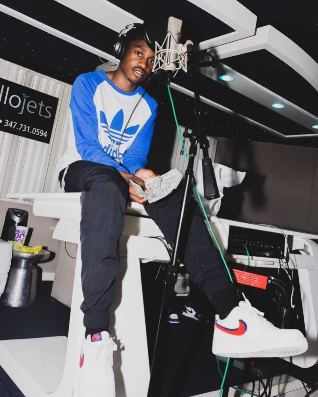 Lil Tjay 桌布 壁紙 — 一位身穿藍白Adidas襯衫的年輕男子隨意地坐在白色工作室桌上，手持現金，戴著耳機，靠近麥克風。