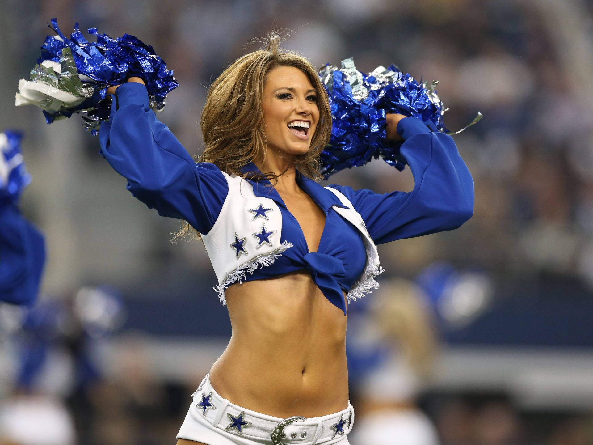 Cheerleader 桌布 壁紙 — 一位身穿藍白相間、帶有星星裝飾的達拉斯牛仔隊啦啦隊員，在體育館內揮舞藍色絨球，笑容燦爛。
