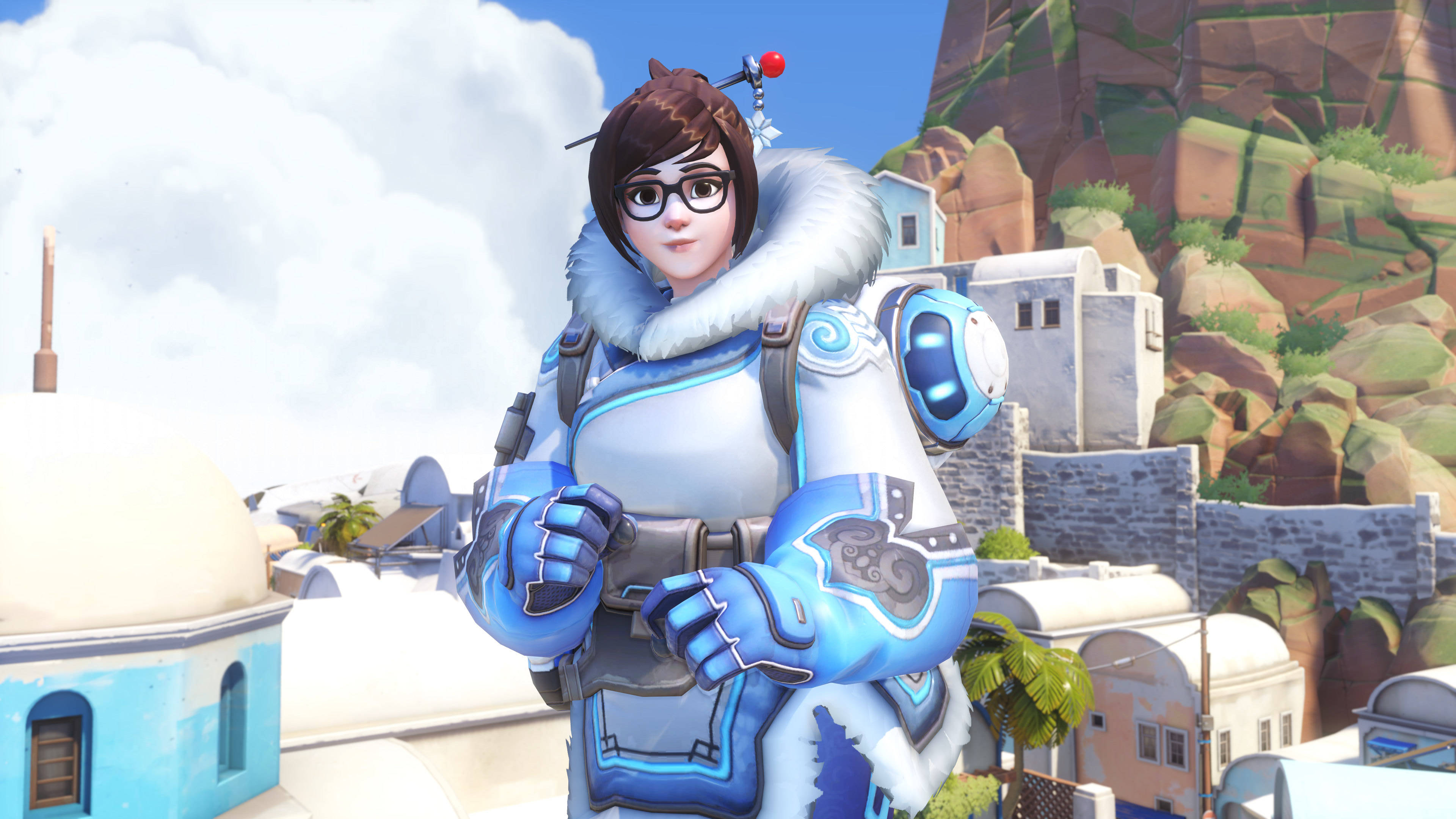 4K 鬥陣特攻 桌布 壁紙 — 一位身穿藍白裝甲服的女性電子遊戲角色自信地站在一個陽光明媚的地中海村莊，村莊有白色圓頂建築和岩石山丘。