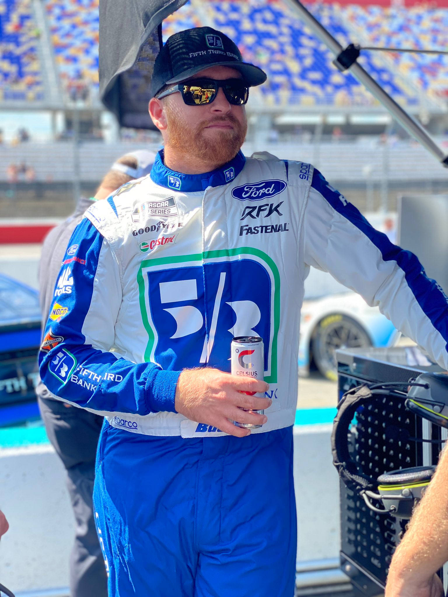 藍色 桌布 壁紙 — 一位身穿藍白賽車服的NASCAR車手，在賽道上的遮陽篷下拿著一個罐子。