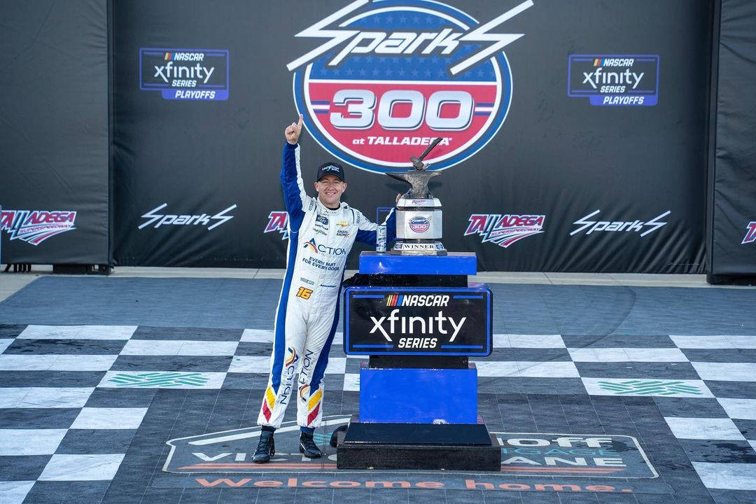 Trophy 桌布 壁紙 — 一位身穿藍白賽車服的NASCAR車手在塔拉迪加超級賽道的方格頒獎台上高舉獎盃慶祝勝利。