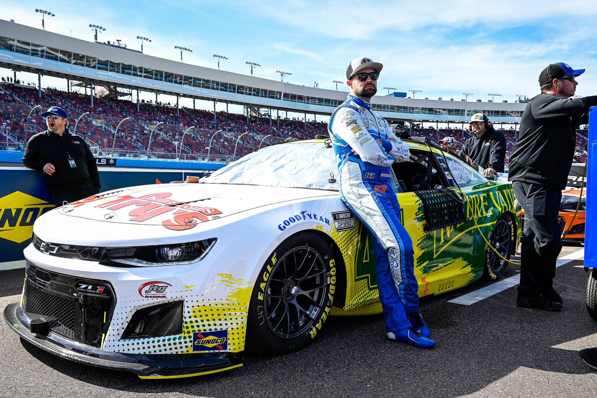 Athlete 桌布 壁紙 — 一位身穿藍白賽車服的NASCAR車手在明亮的日光下，於擠滿觀眾的賽道上靠在他鮮豔的黃綠色賽車上。