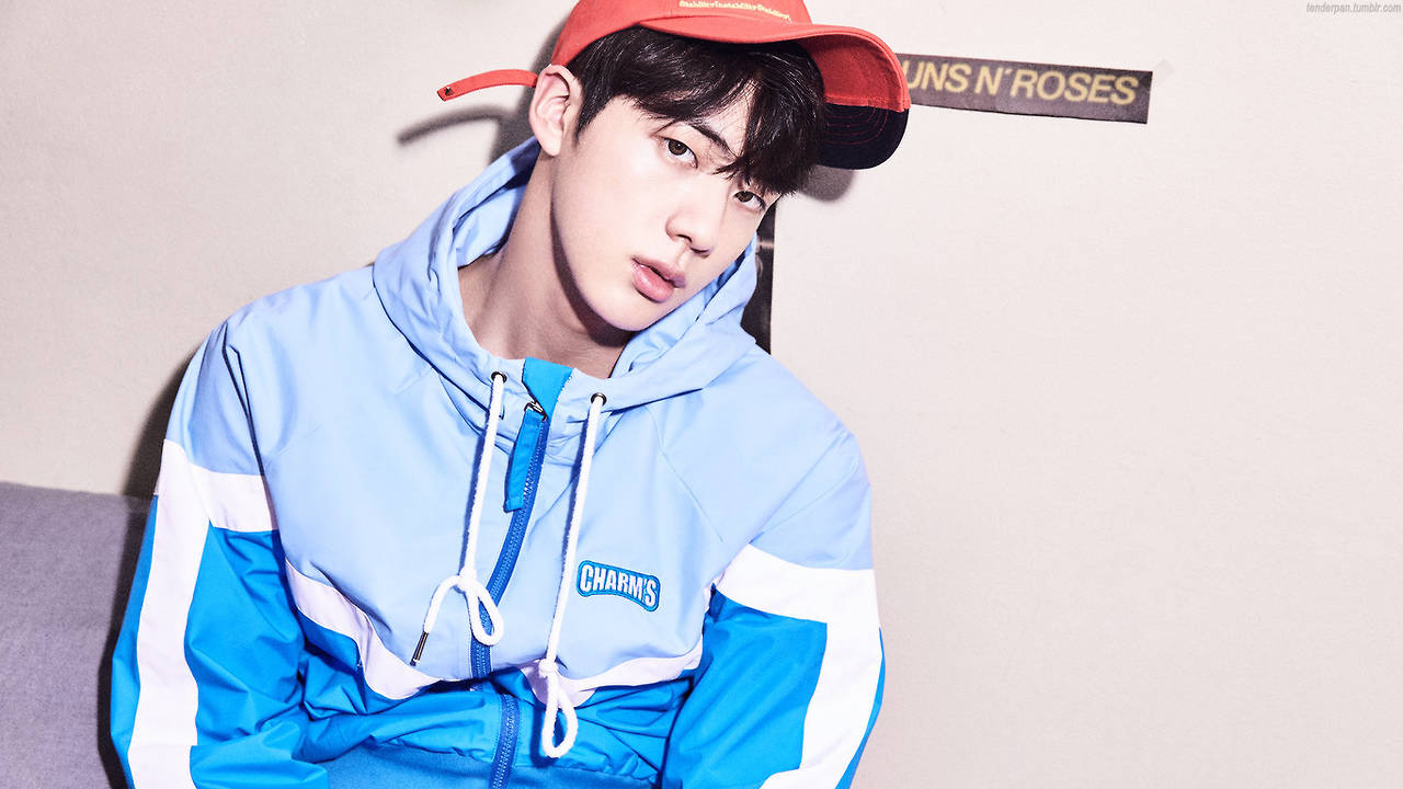 kim seok jin 桌布 壁紙 — 一位身穿藍白運動外套和紅色帽子的年輕男子，在一個帶有「GUNS N' ROSES」標誌的素色牆前擺姿勢。