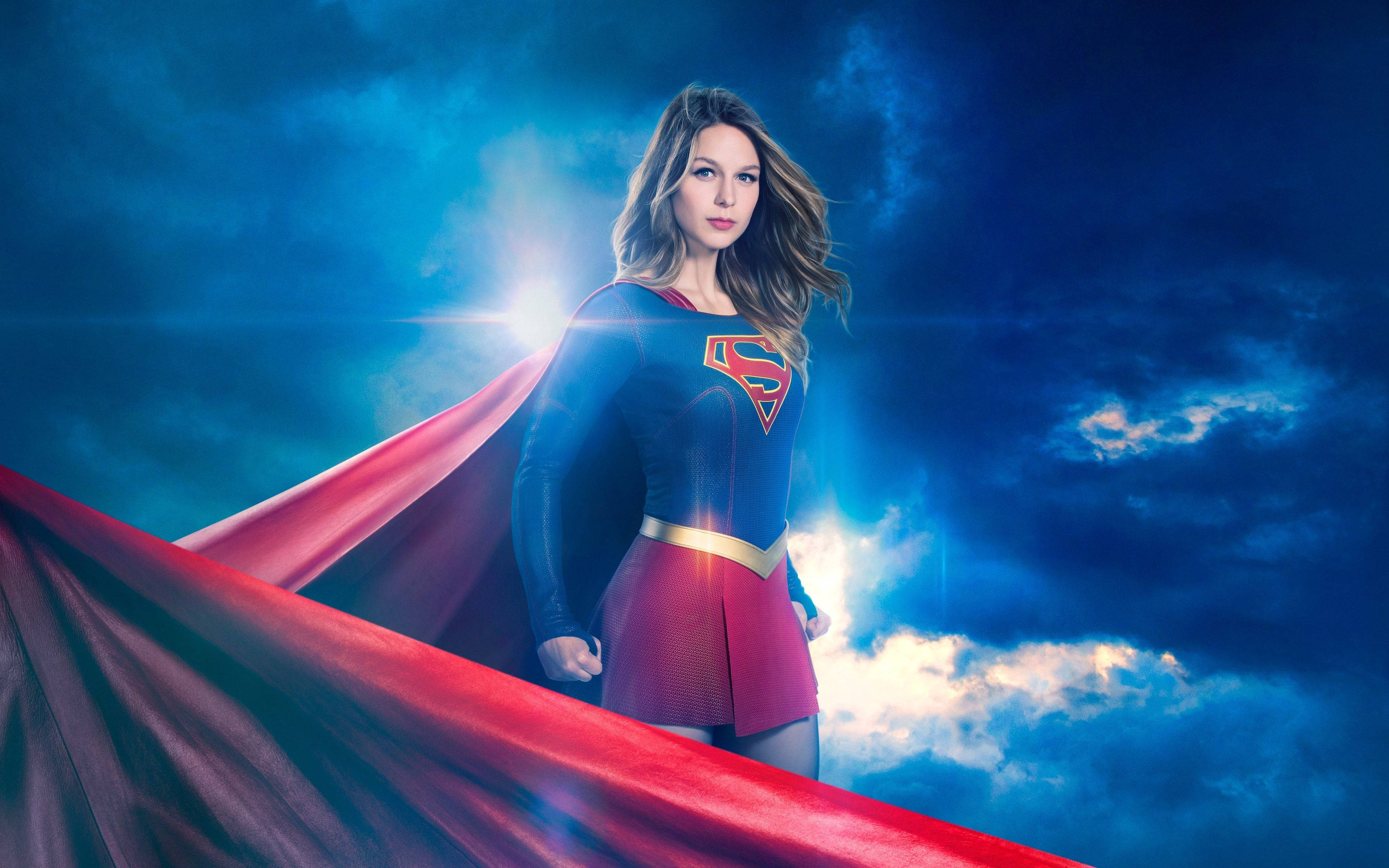 Supergirl 桌布 壁紙 — 一位身穿藍紅服裝的女超級英雄自信地站在戲劇性的多雲天空前，背後有燦爛的陽光爆發。