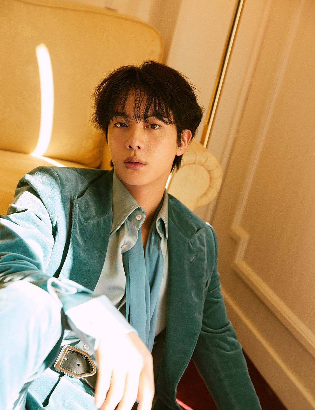 kim seok jin 桌布 壁紙 — 一位身穿藍綠色天鵝絨西裝外套的年輕男子坐在金色椅子上，沐浴在從窗戶灑進的溫暖陽光中。