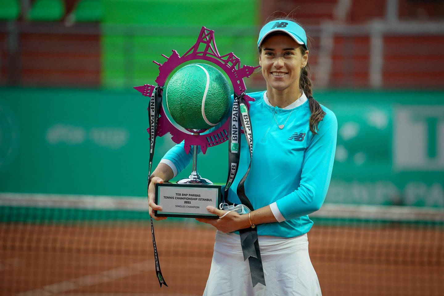 Tennis 桌布 壁紙 — 一位身穿藍綠色服裝、面帶微笑的女網球選手，在紅土錦標賽中手持一個大型綠色網球獎盃。