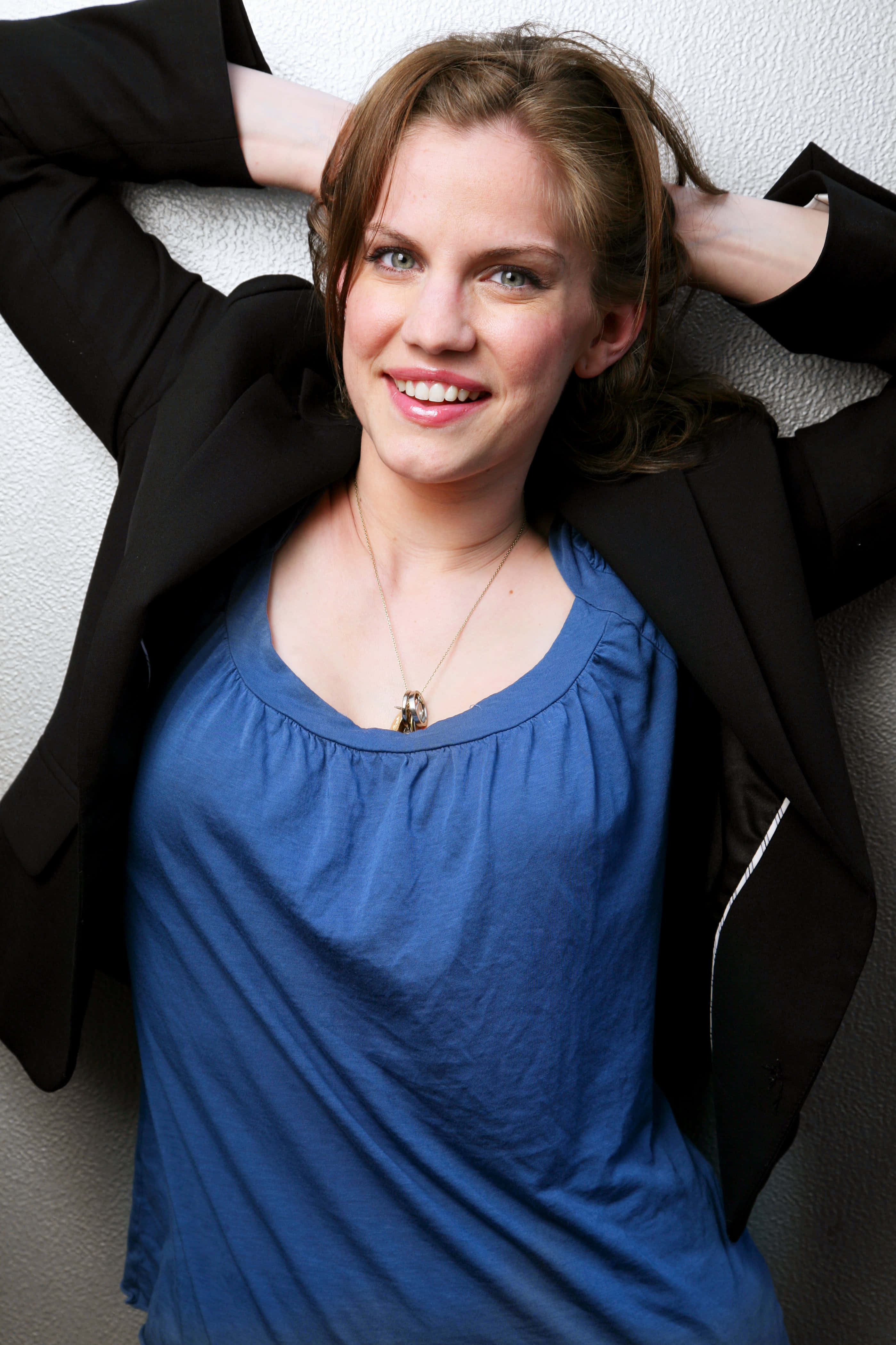 anna chlumsky 桌布 壁紙 — 一位身穿藍色上衣和黑色西裝外套的女士微笑著，雙手放在腦後，靠在一面有紋理的牆上。