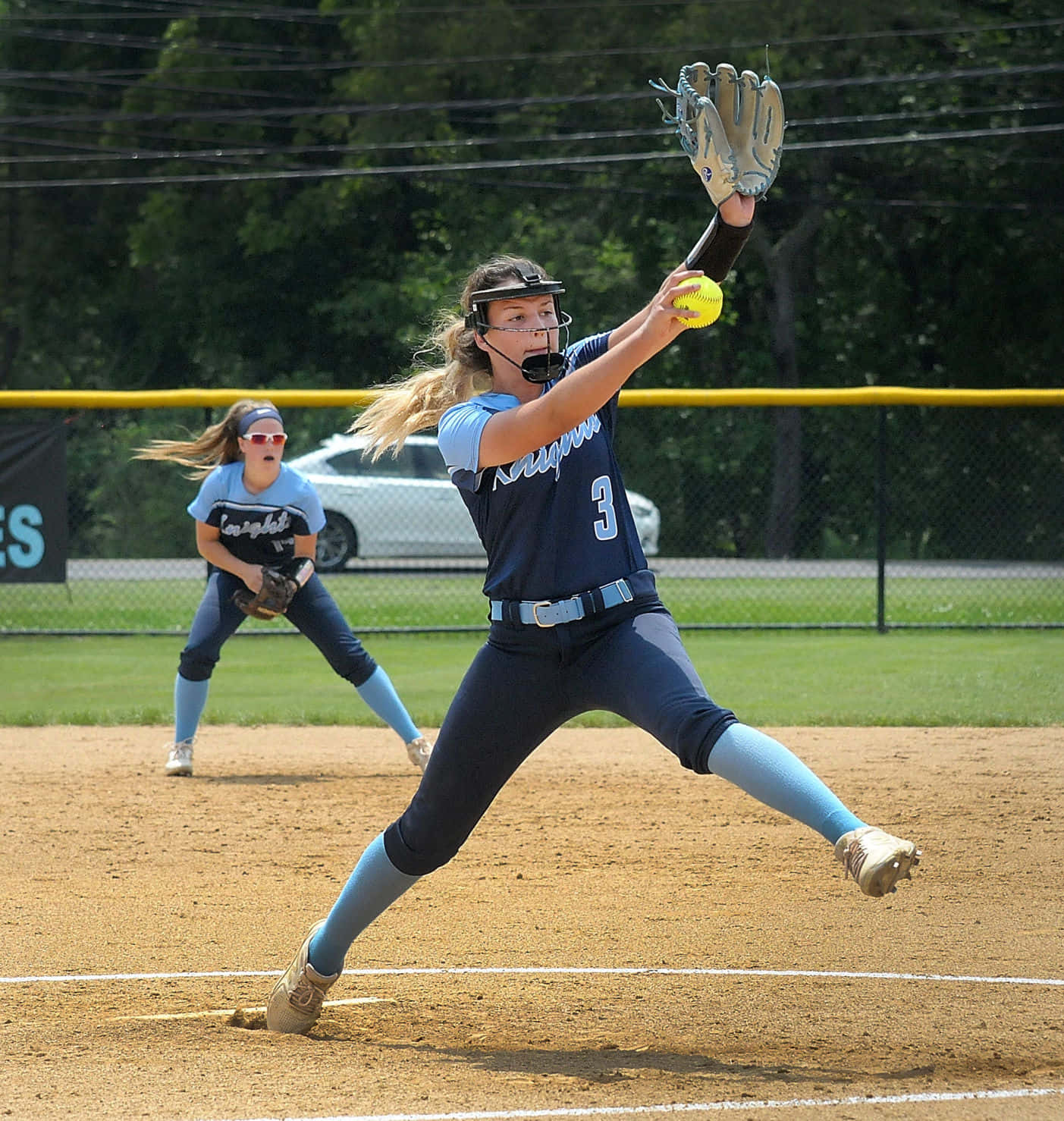 Softball 桌布 壁紙 — 一位身穿藍色制服的女壘球投手在陽光明媚的球場上準備投出黃色球，背景有隊友。