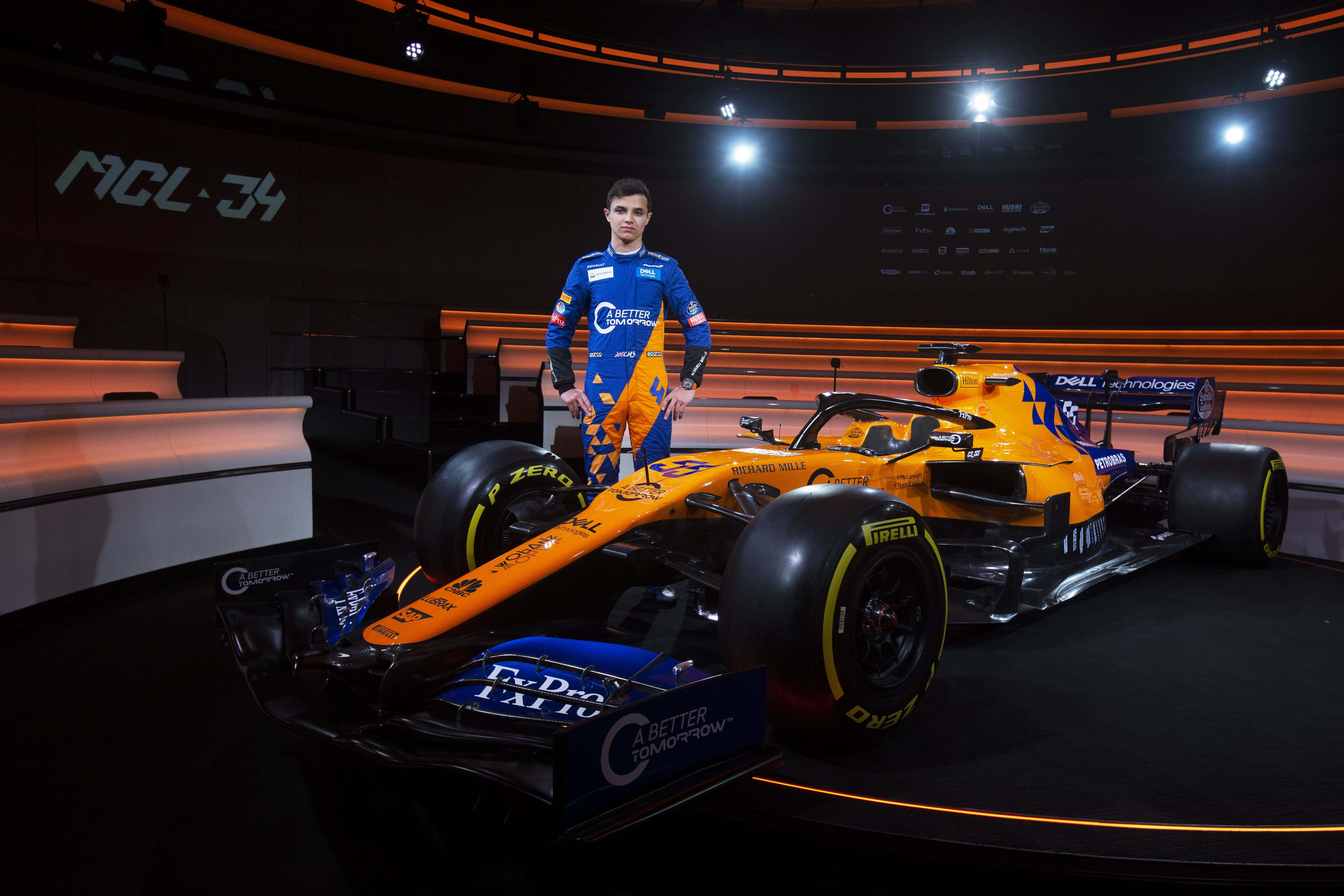 lando norris 桌布 壁紙 — 一位身穿藍色和橙色賽車裝備的F1車手，在明亮的攝影棚燈光下自信地站在他充滿活力的橙色和藍色MCL34賽車旁。