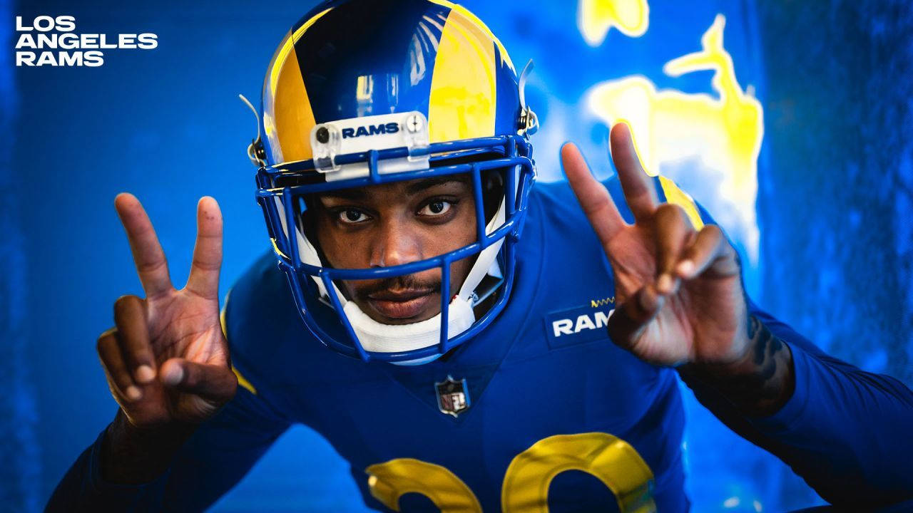Ramsey 桌布 壁紙 — 一位身穿藍色和金色裝備的洛杉磯公羊隊美式足球員在發光的藍色背景前做出和平手勢。