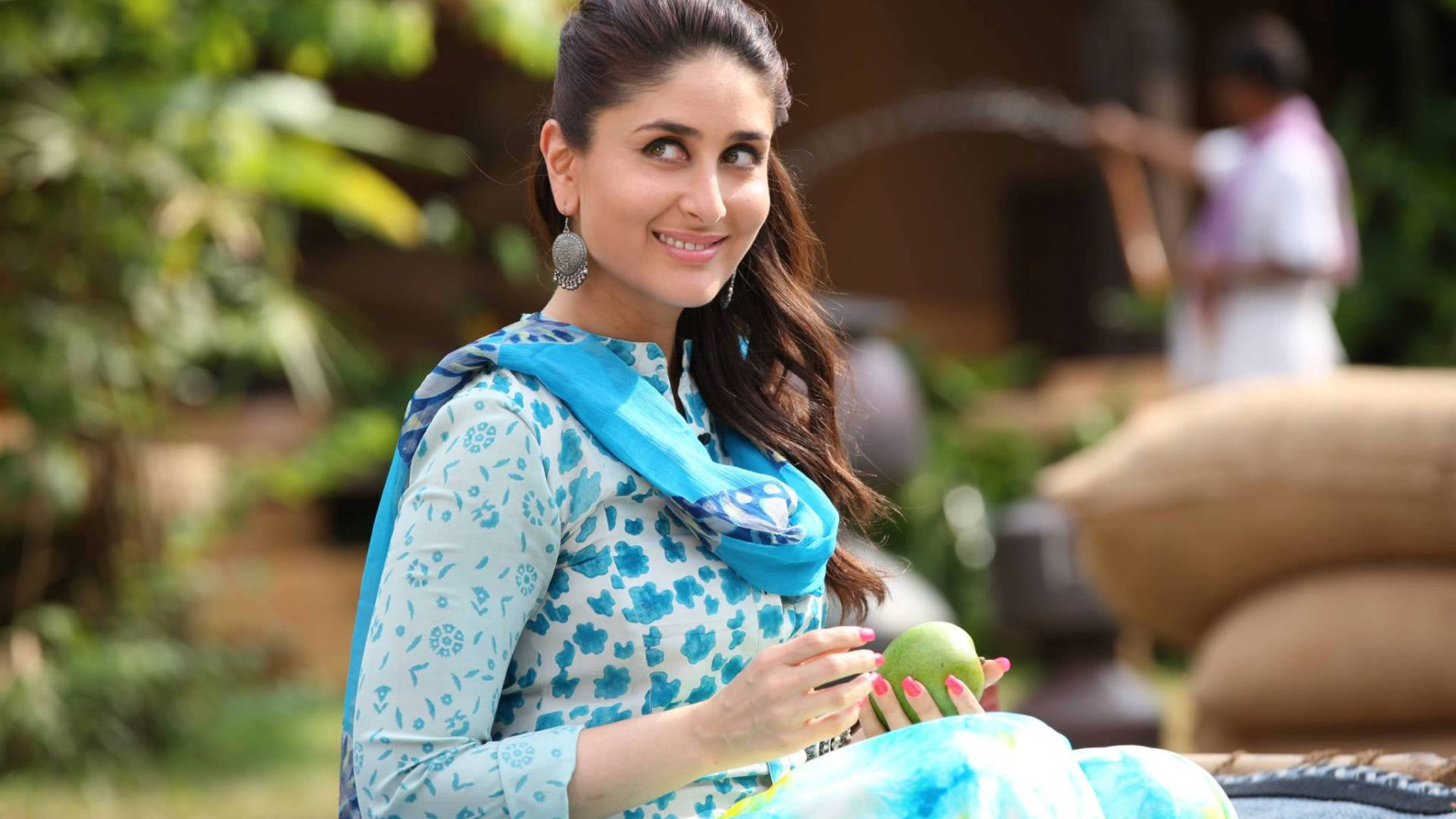 Kareena 桌布 壁紙 — 一位身穿藍色圖案服裝的女性在茂盛的花園中微笑著手持綠色芒果。