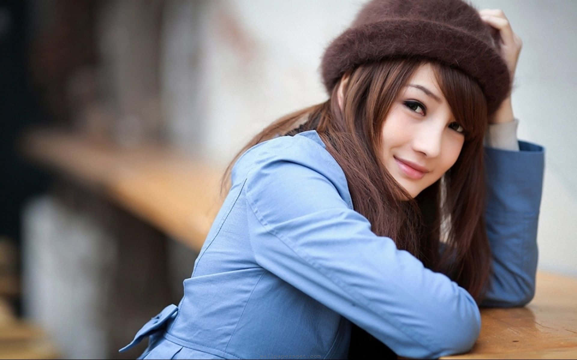 美女 桌布 壁紙 — 一位身穿藍色外套、戴著棕色帽子的年輕女性，在舒適的室內環境中靠在木桌上溫柔微笑。