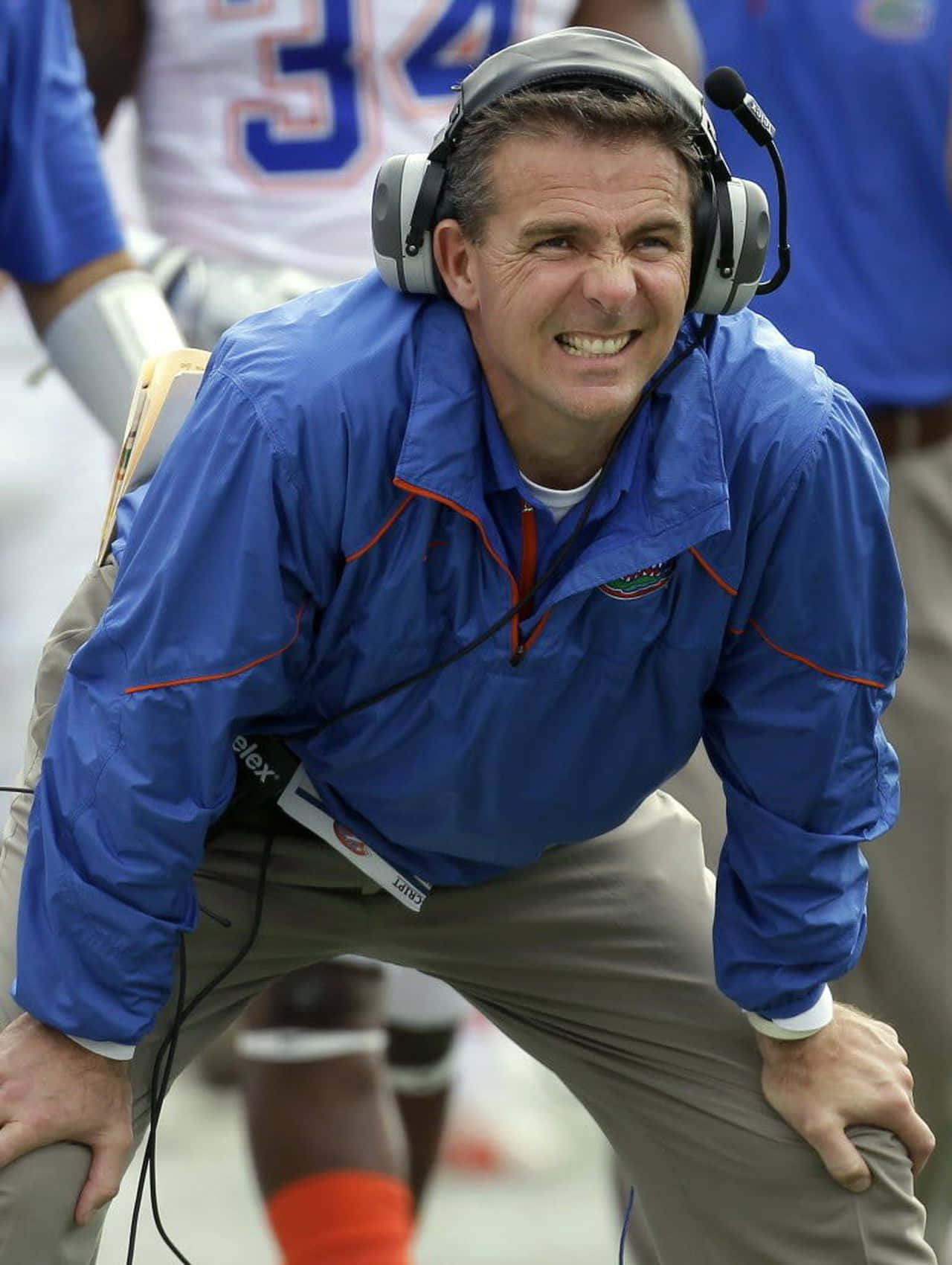 Urban Meyer 桌布 壁紙 — 一位身穿藍色外套、戴著耳機的美式足球教練在比賽期間於邊線向前傾，露出強烈的笑容。