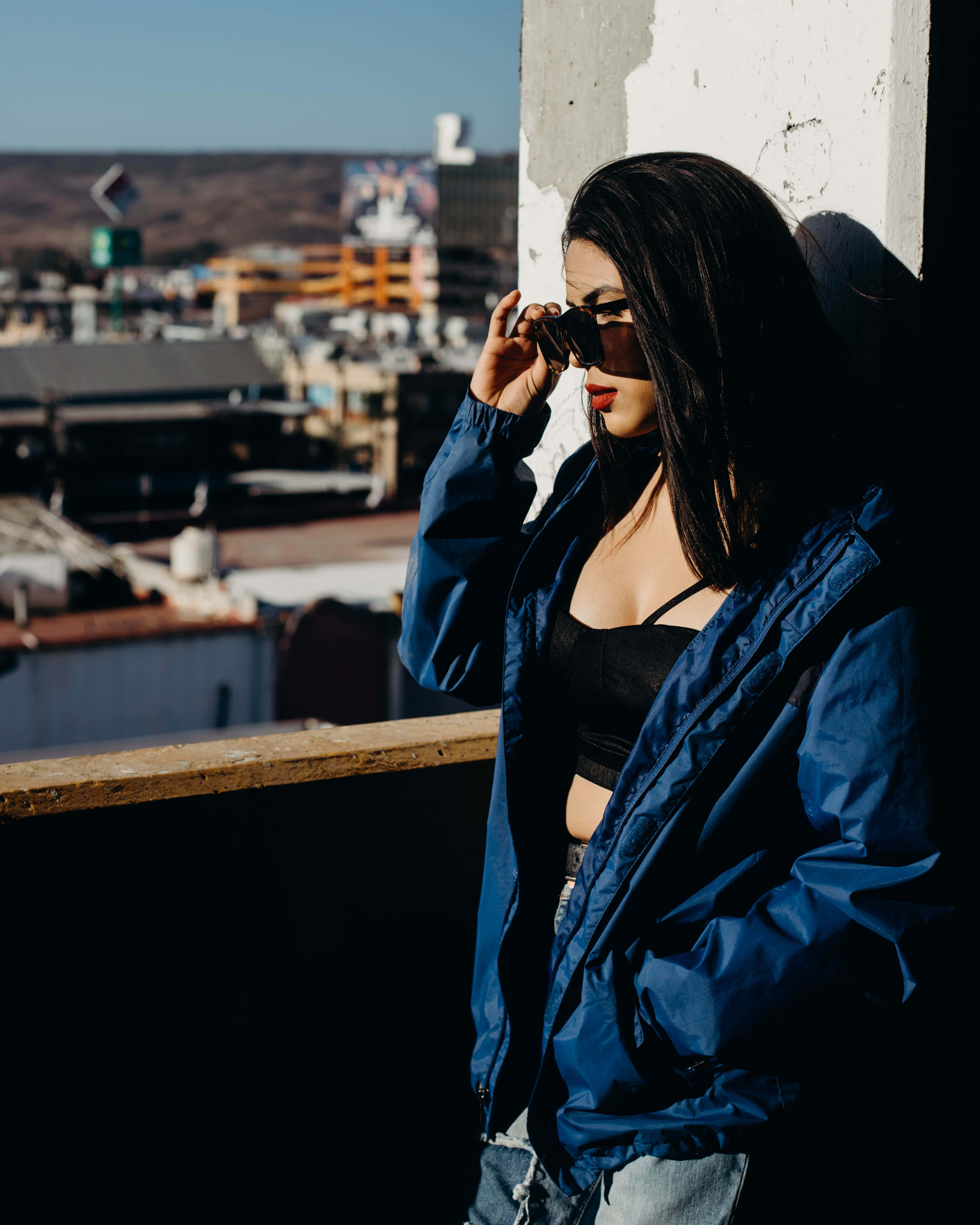 Navy Blue Jacket 桌布 壁紙 — 一位身穿藍色外套的女性靠在混凝土柱上調整太陽眼鏡，身處陽光普照的屋頂，俯瞰城市景觀。