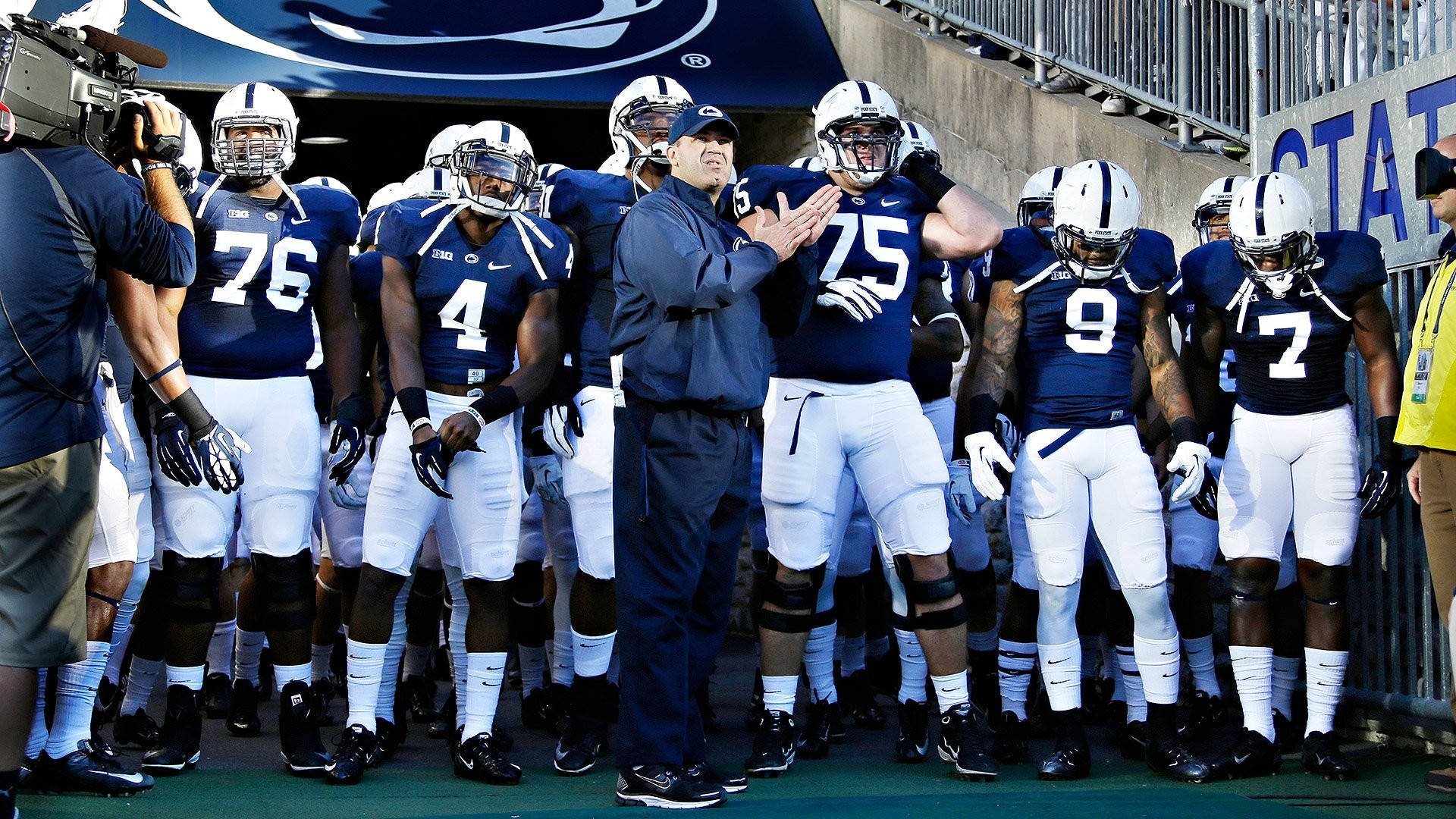 Penn State 桌布 壁紙 — 一位身穿藍色外套的足球教練在鼓掌，他的球隊穿著藍白相間的球衣在邊線準備就緒。