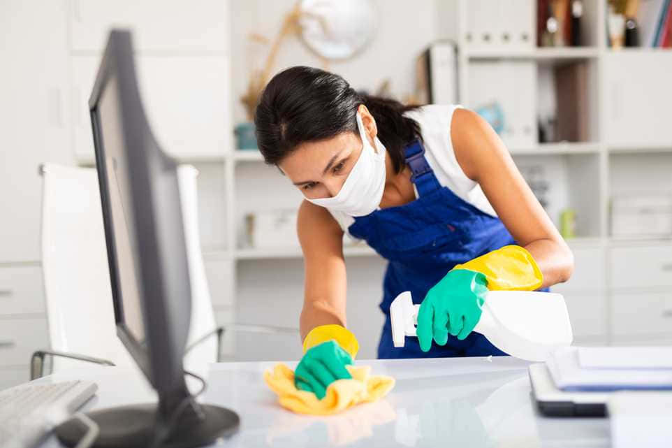 Cleaning 桌布 壁紙 — 一位身穿藍色工裝褲和口罩的女性，在明亮的現代辦公室裡用黃色布仔細擦拭電腦顯示器。