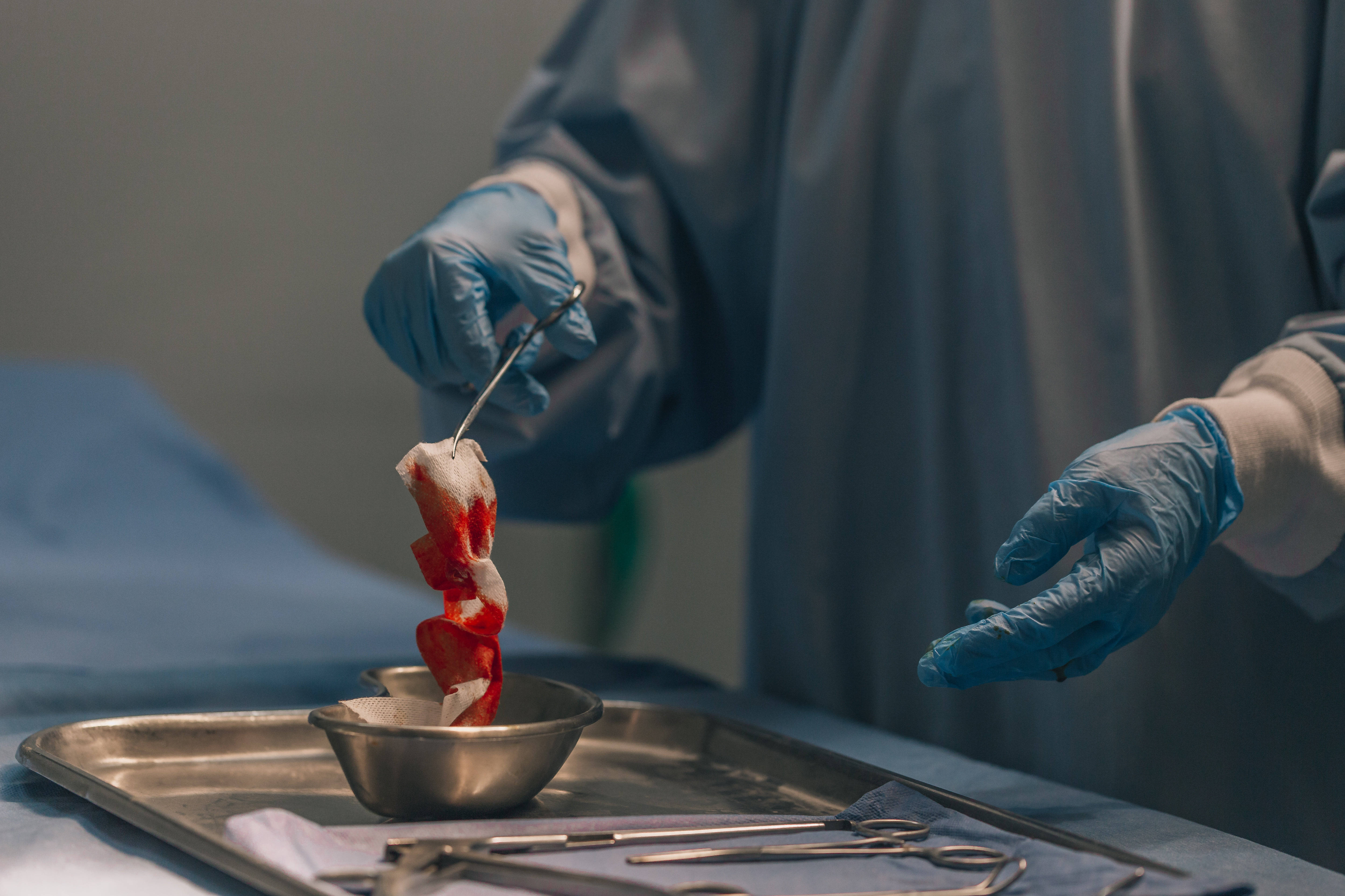 Doctors 桌布 壁紙 — 一位身穿藍色手術服和手套的外科醫生用鑷子在金屬手術托盤上處理帶血的組織樣本。