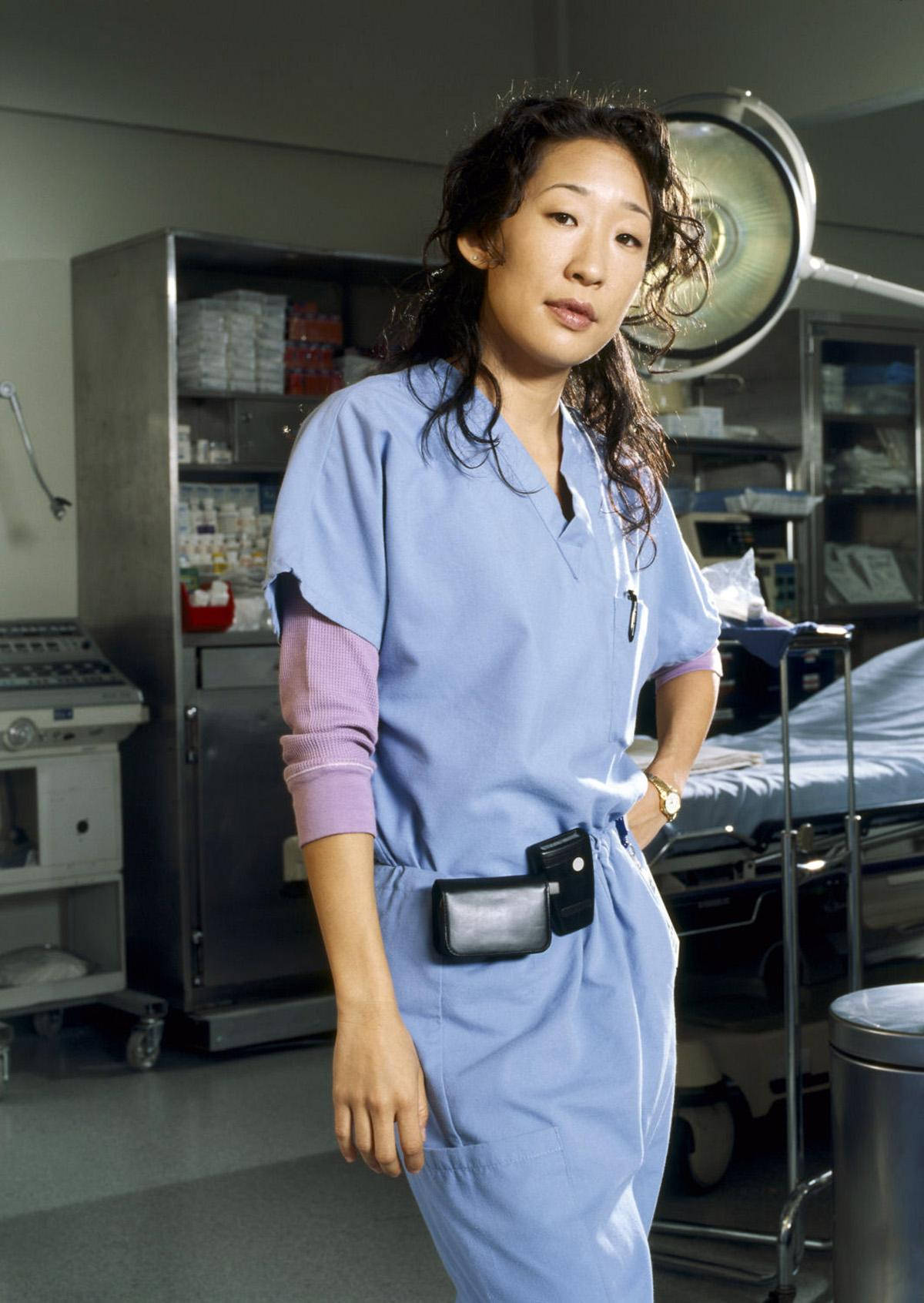 greys anatomy 桌布 壁紙 — 一位身穿藍色手術服的女外科醫生自信地站在光線充足的手術室中，頭頂有醫療設備和手術燈。