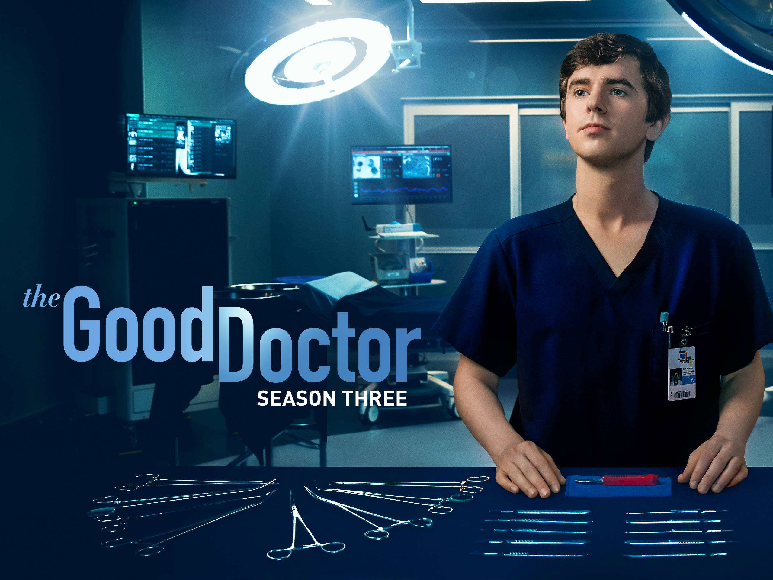Freddie Highmore 桌布 壁紙 — 一位身穿藍色手術服的年輕醫生站在光線昏暗的手術室中，頭頂明亮的燈光，面前擺放著醫療工具。