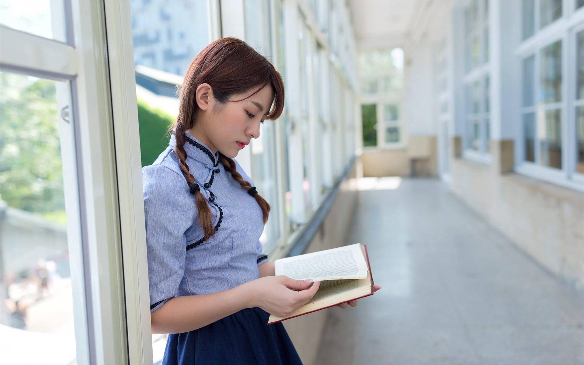 Reading 桌布 壁紙 — 一位身穿藍色校服的年輕女子在明亮的學校走廊裡靠著窗戶看書。