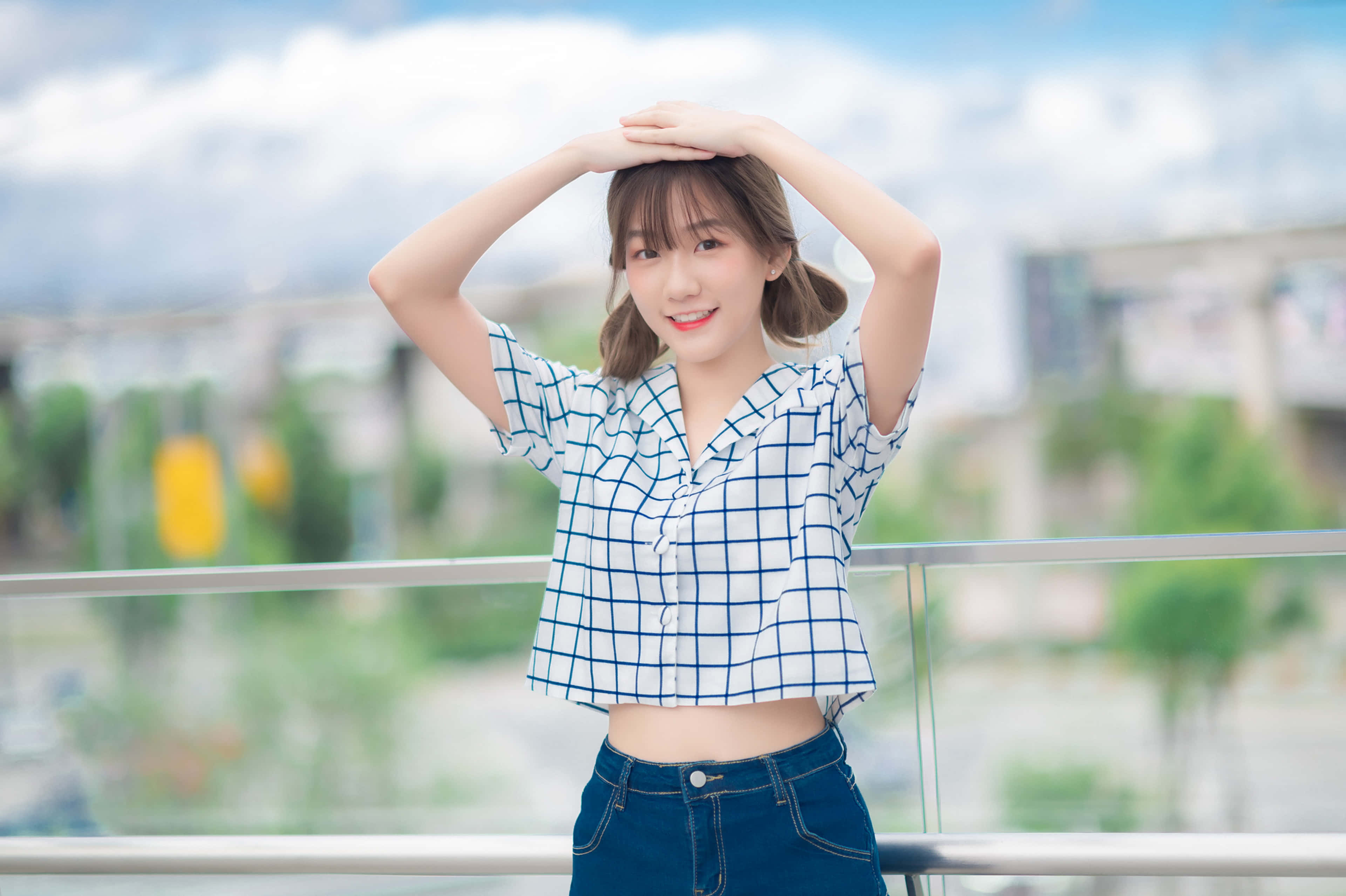 女孩 桌布 壁紙 — 一位身穿藍色格子襯衫的開朗年輕女性，在陽光明媚的陽台上舉起雙手放在頭頂上微笑擺姿勢。