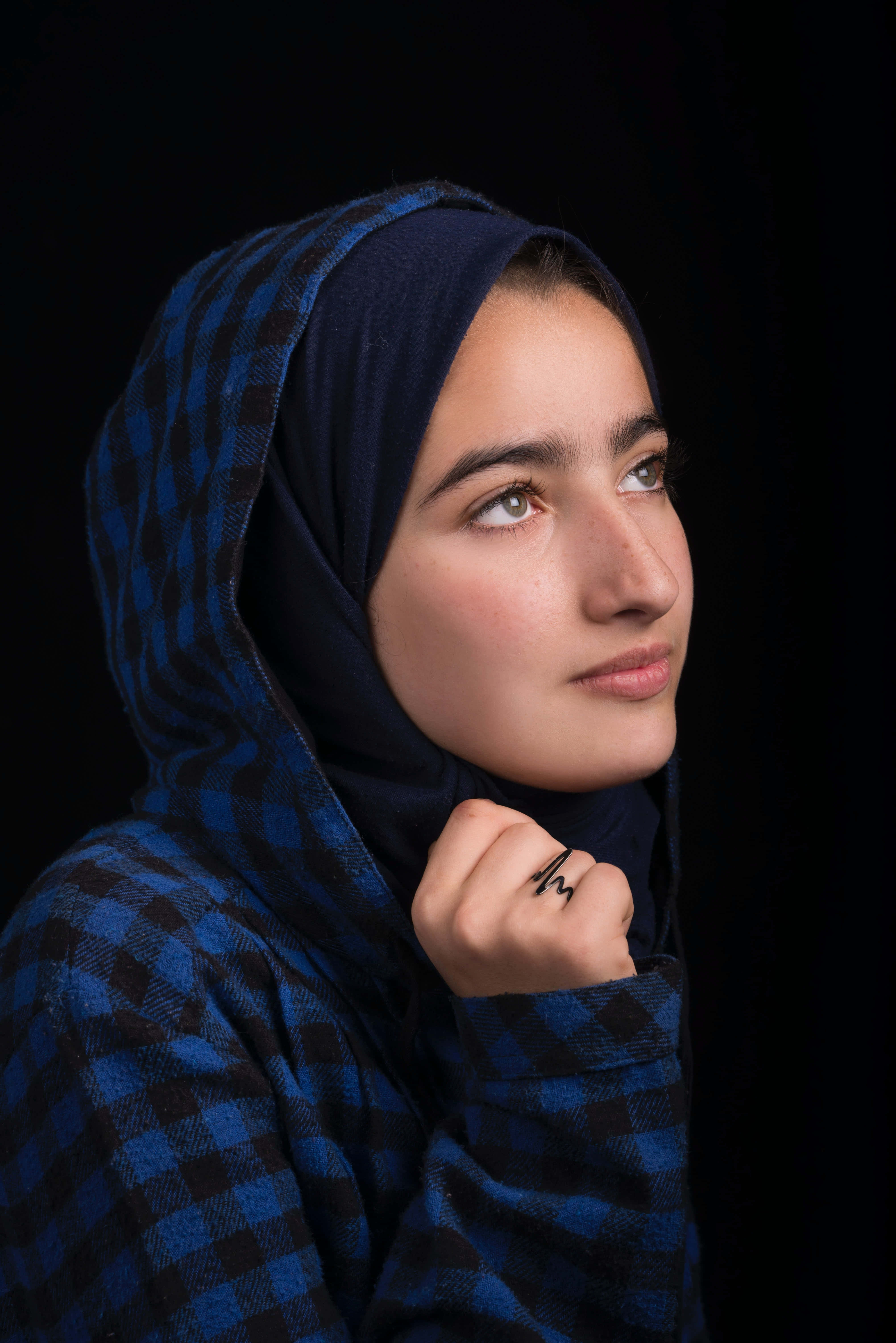 Navy Blue Scarf 桌布 壁紙 — 一位身穿藍色格紋希賈布的年輕女子，在純黑背景下若有所思地向上凝視。