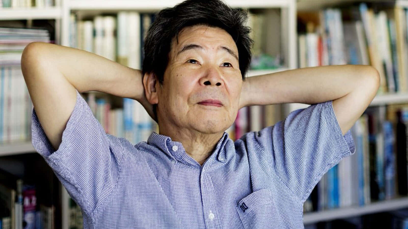 Isao Takahata 桌布 壁紙 — 一位身穿藍色格紋襯衫的長者，在擺滿書架的房間裡將雙臂枕在腦後休息。