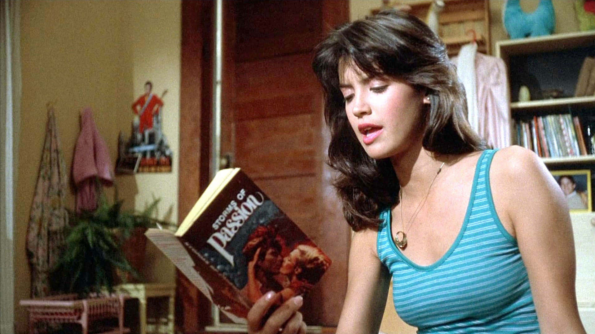 Phoebe Cates 桌布 壁紙 — 一位身穿藍色條紋背心的女性，在溫暖照明的室內房間裡，閱讀一本名為《Stronger of Passion》的書，房間內有書架和裝飾。