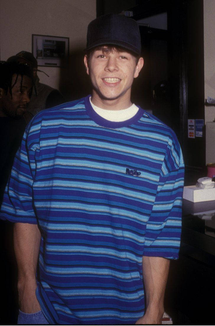 Mark Wahlberg 桌布 壁紙 — 一位身穿藍色條紋襯衫、戴黑色帽子的年輕男子在室內溫暖的人造燈光下燦爛地微笑。