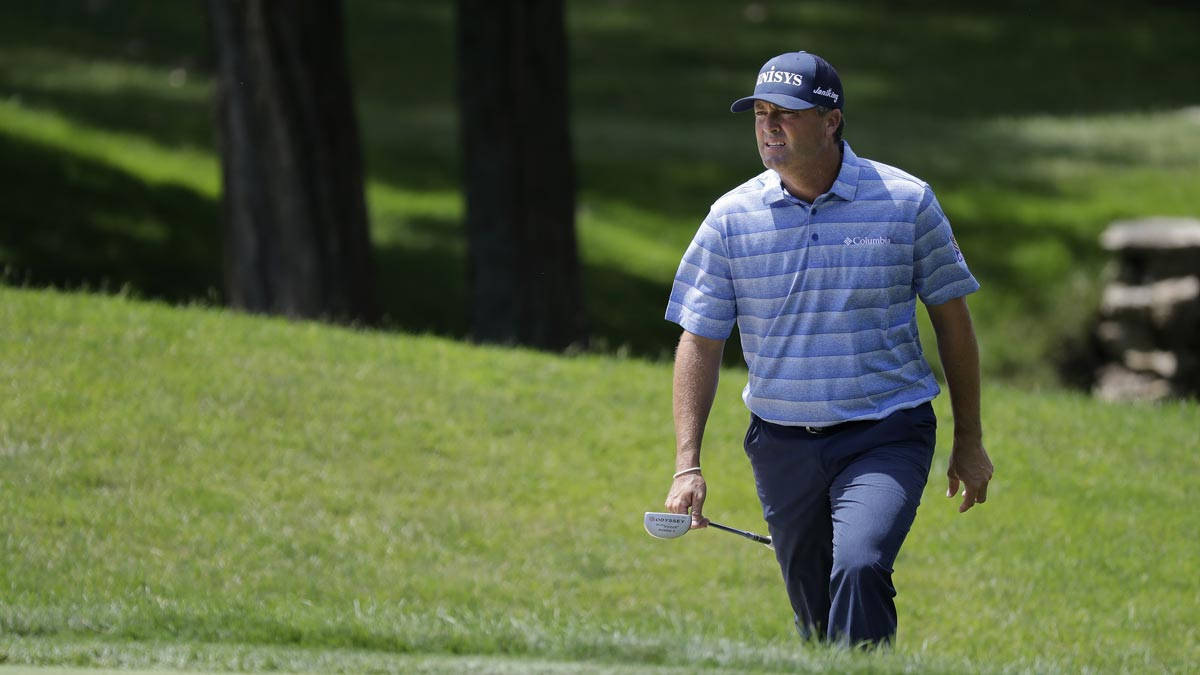 Ryan Palmer 桌布 壁紙 — 一位身穿藍色條紋襯衫的高爾夫球手，手持球桿，走過陽光明媚的綠色球道，背景有樹木。