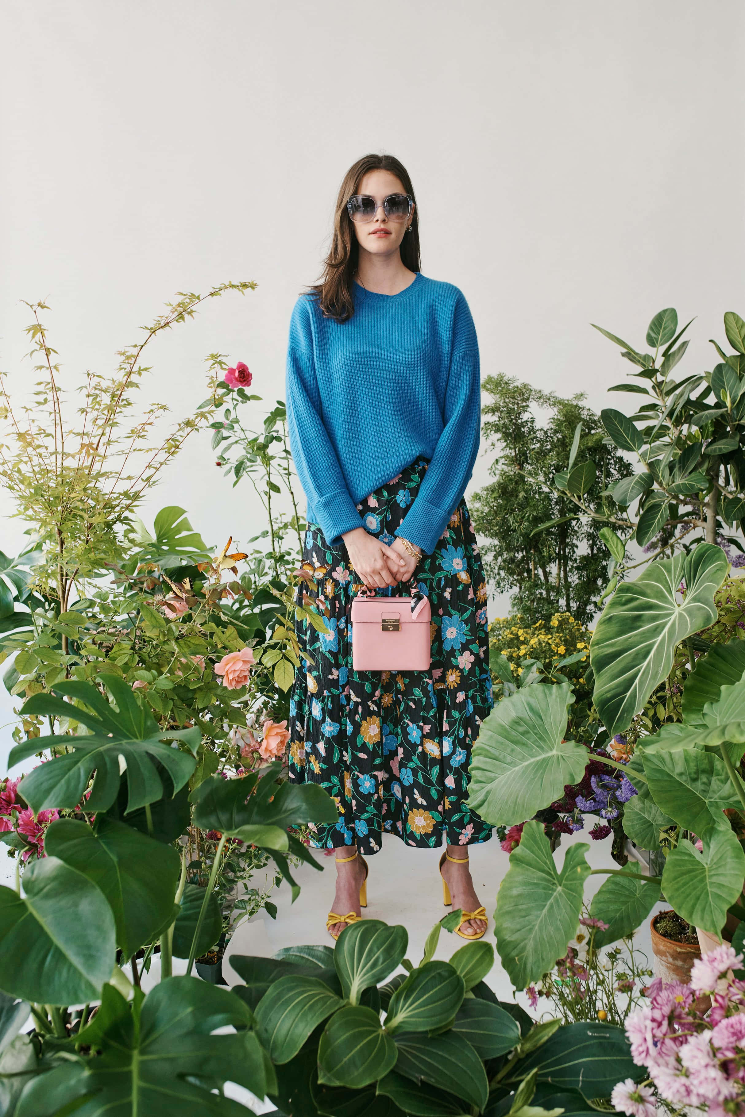 kate spade 桌布 壁紙 — 一位身穿藍色毛衣和花卉裙的女性，被茂盛的綠色植物和色彩繽紛的花朵環繞。