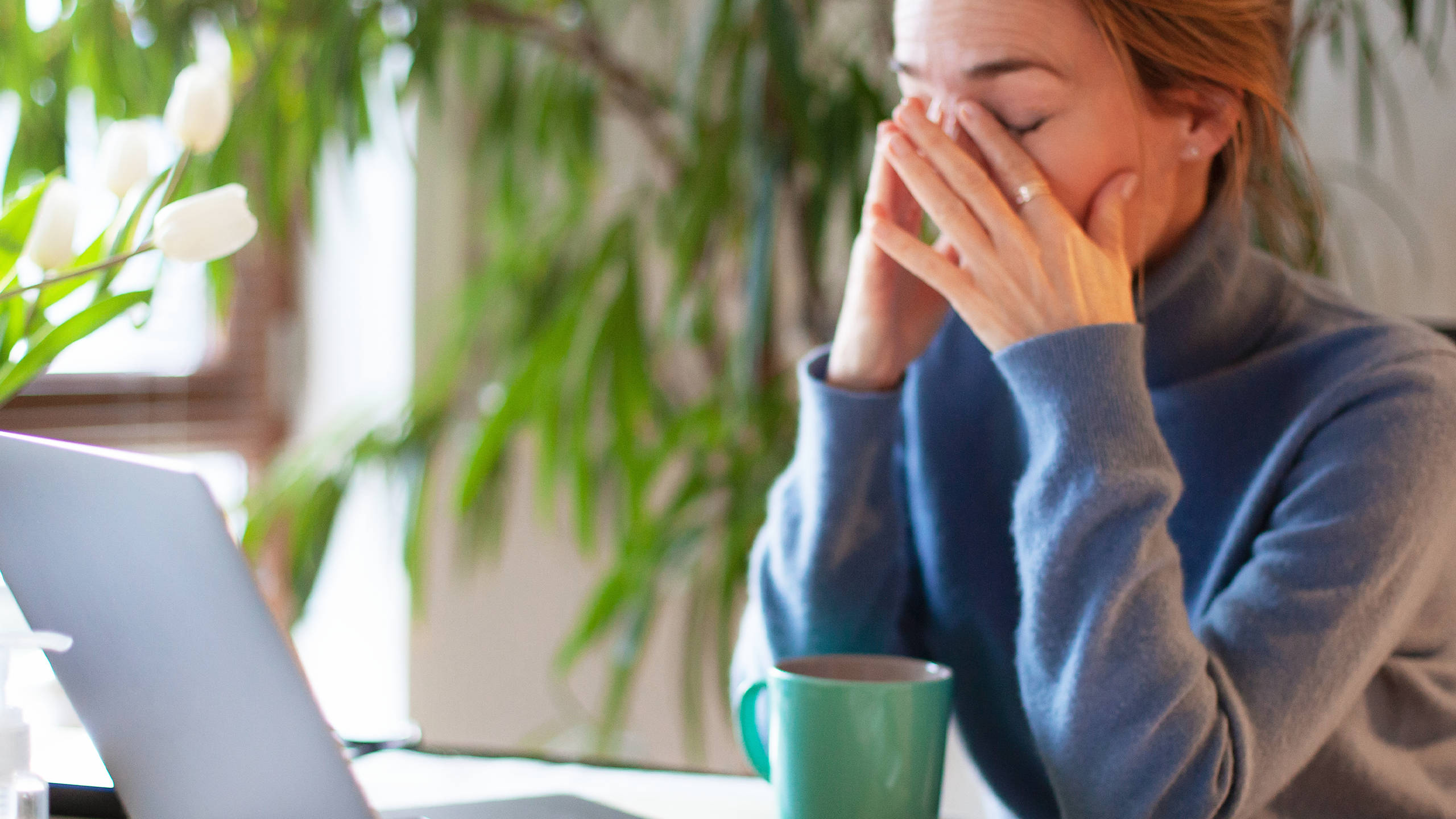 tired 桌布 壁紙 — 一位身穿藍色毛衣的女性坐在辦公桌前，桌上放著筆記型電腦和一個綠色杯子，她用手遮住了臉。