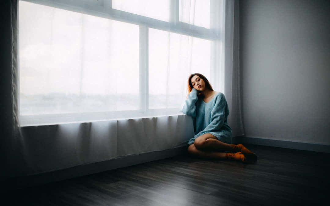 Woman 桌布 壁紙 — 一位身穿藍色毛衣的女性靜靜地坐在木地板上，旁邊是一扇帶有薄紗窗簾的大窗戶，沐浴在柔和自然光中。