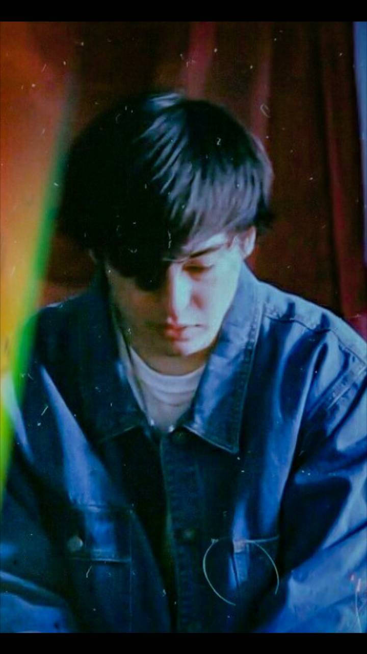 Joji 桌布 壁紙 — 一位身穿藍色牛仔外套的年輕男子，在溫暖色調、柔和模糊的背景前，帶著沉思的表情向下凝視。