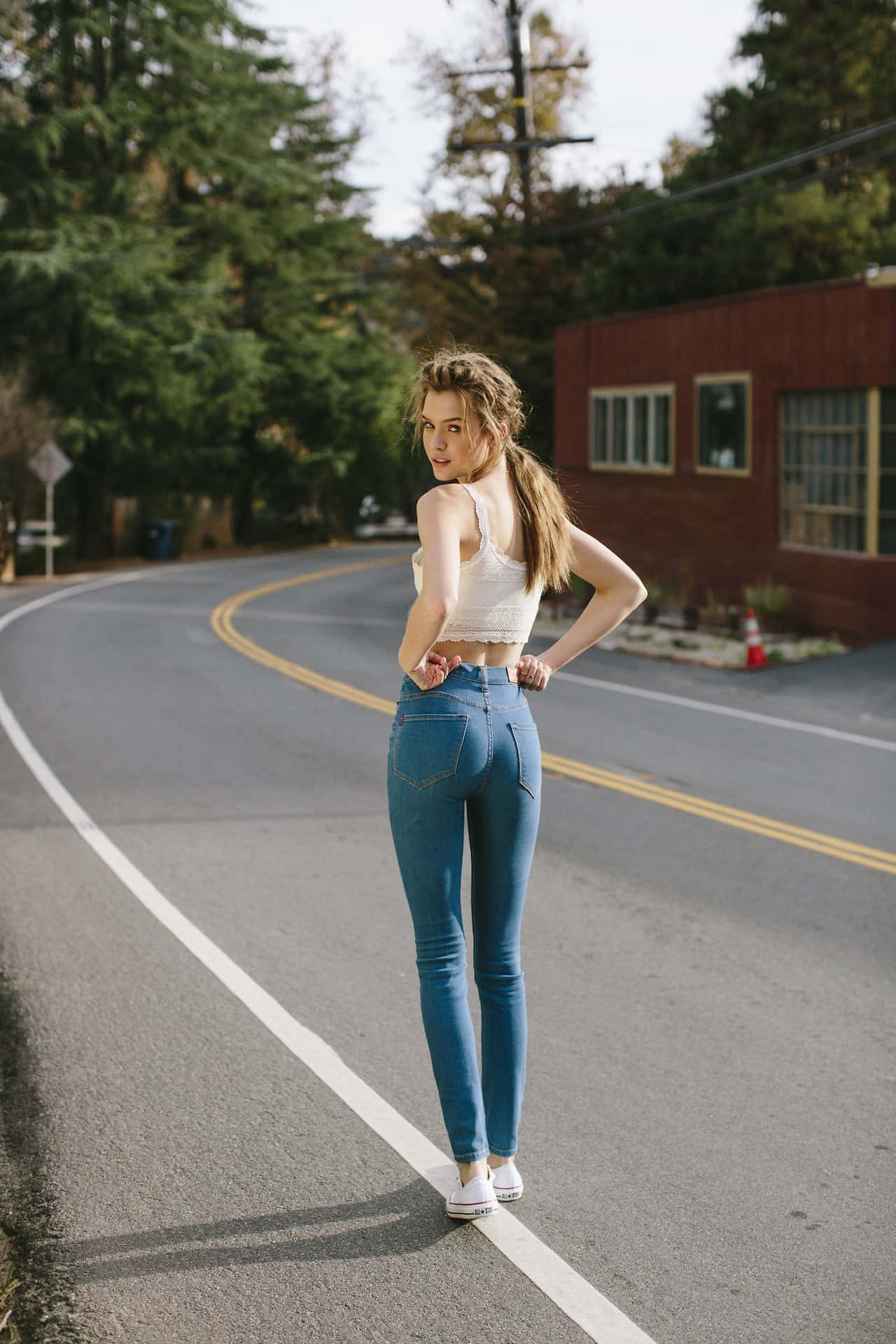 Beautiful Girls 桌布 壁紙 — 一位身穿藍色牛仔褲和白色上衣的年輕女性自信地站在一條彎曲的道路上，背景有樹木和一棟紅色建築。