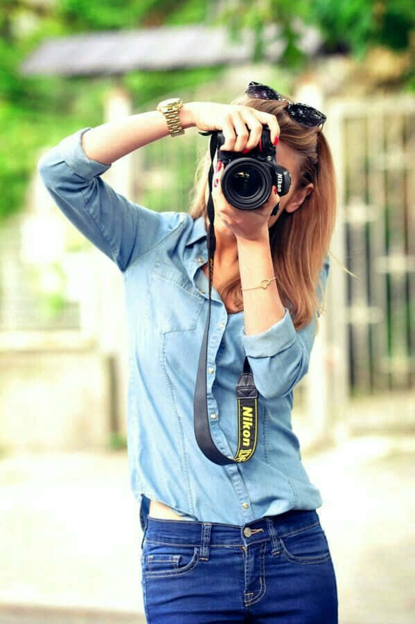 Holding Camera 桌布 壁紙 — 一位身穿藍色牛仔襯衫和牛仔褲的年輕女性，在戶外綠意盎然的背景下，將Nikon相機舉到臉前。