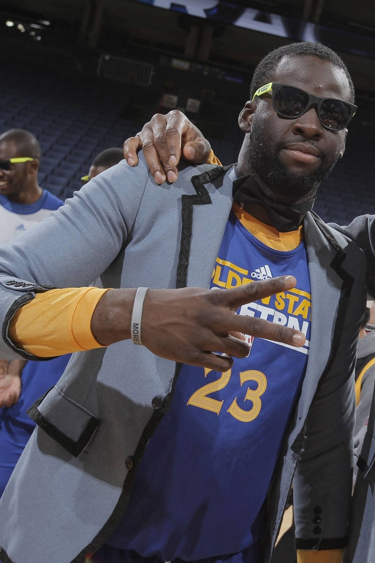 Draymond Green 桌布 壁紙 — 一位身穿藍色球衣和灰色西裝外套的籃球運動員，在室內戴著太陽眼鏡比出和平手勢。