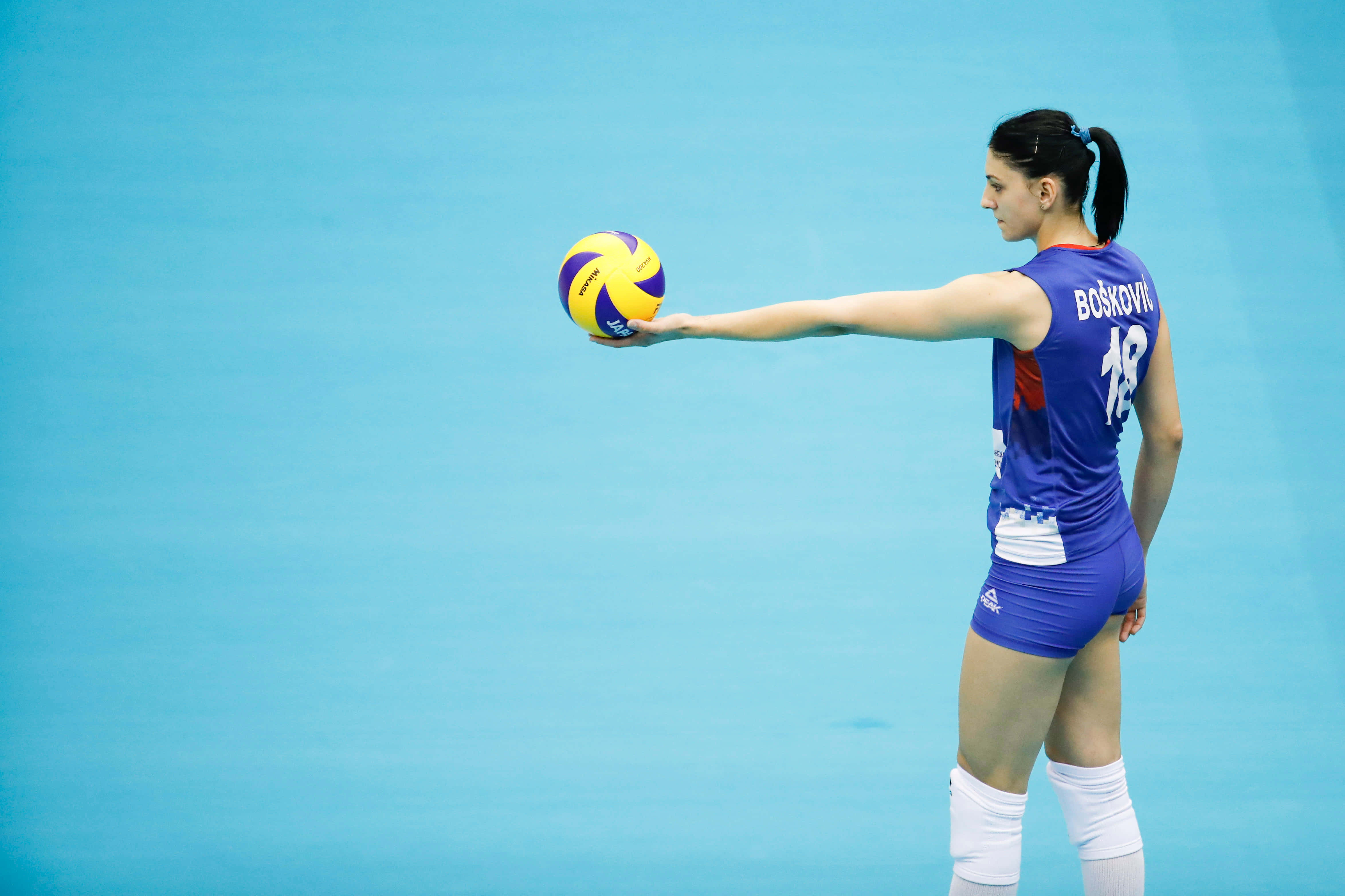 Volleyball 桌布 壁紙 — 一位身穿藍色球衣的女排球運動員，在純藍色球場背景下伸出手臂，手持一個黃紫相間的球。