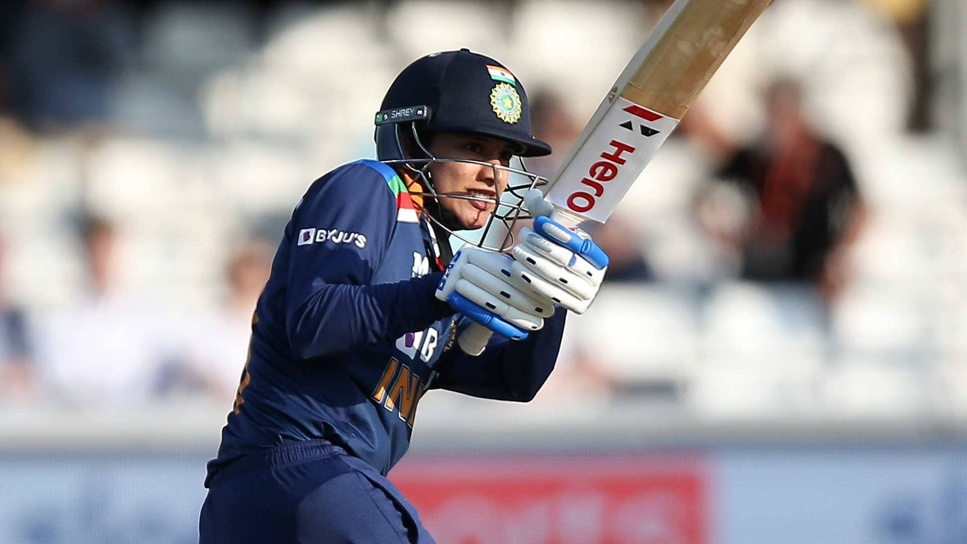 Smriti Mandhana 桌布 壁紙 — 一位身穿藍色球衣的女板球運動員在日間比賽中揮動球棒，背景的體育館座位模糊不清。