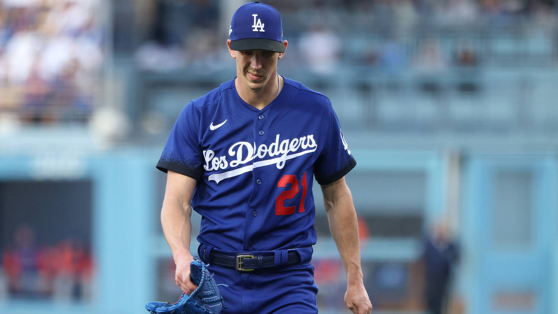 walker buehler 桌布 壁紙 — 一位身穿藍色球衣的洛杉磯道奇棒球選手，在陽光明媚的球場上拿著手套行走。